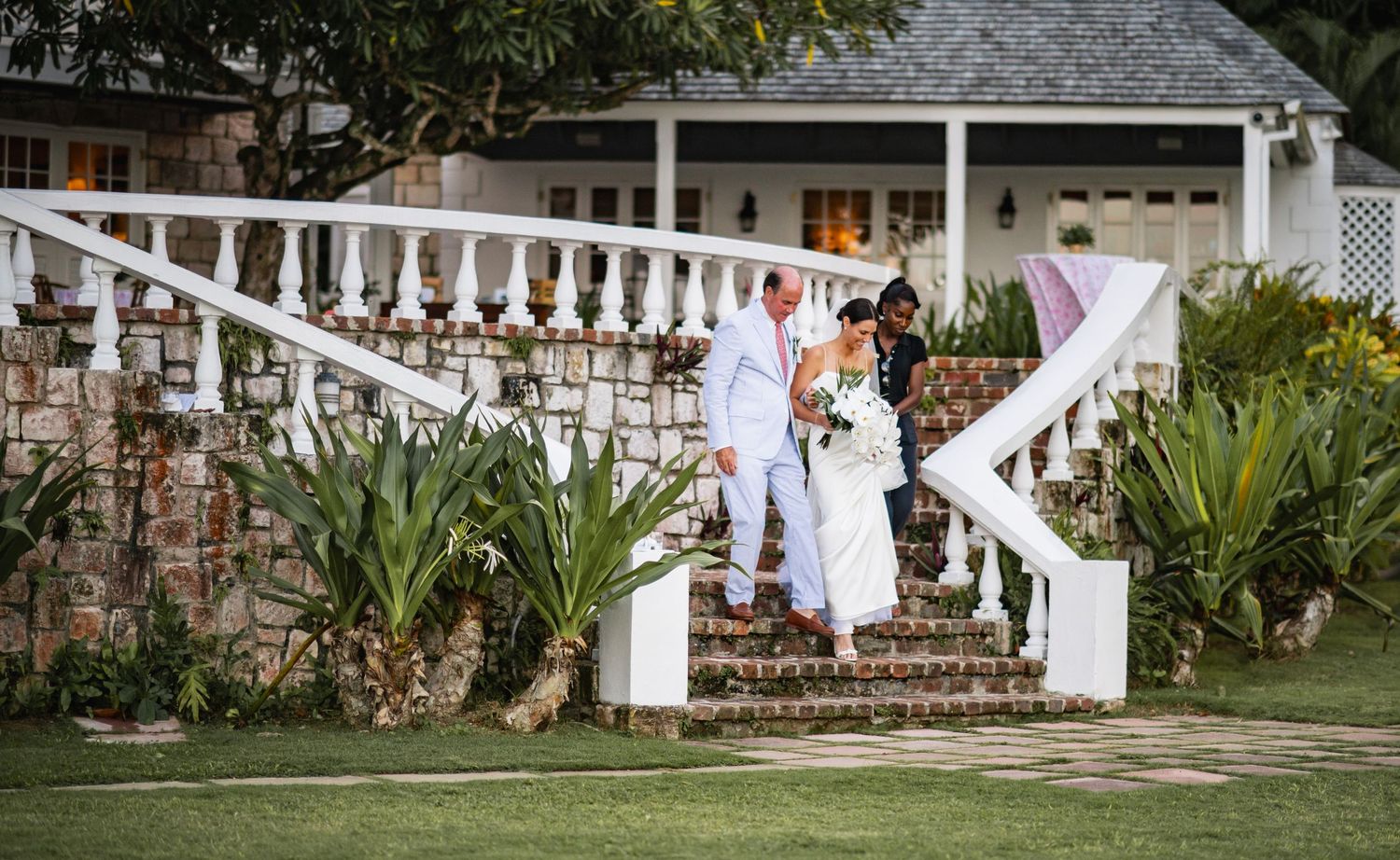 A couple stands together on brick steps leading to a white railed porch of a charming house with tropical landscaping.