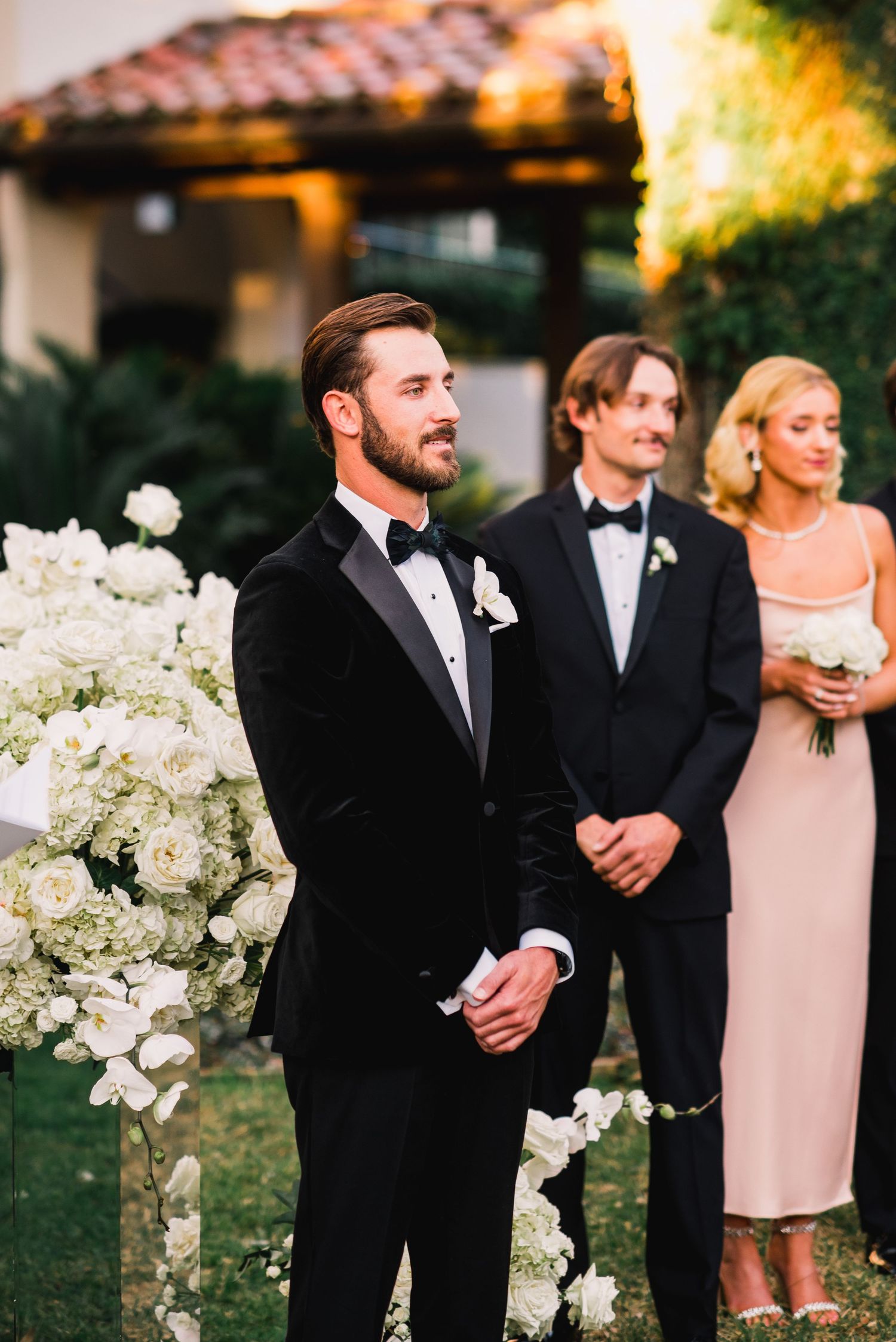 Wedding party members in black tuxedos and blush pink dresses stand together during an outdoor evening ceremony.