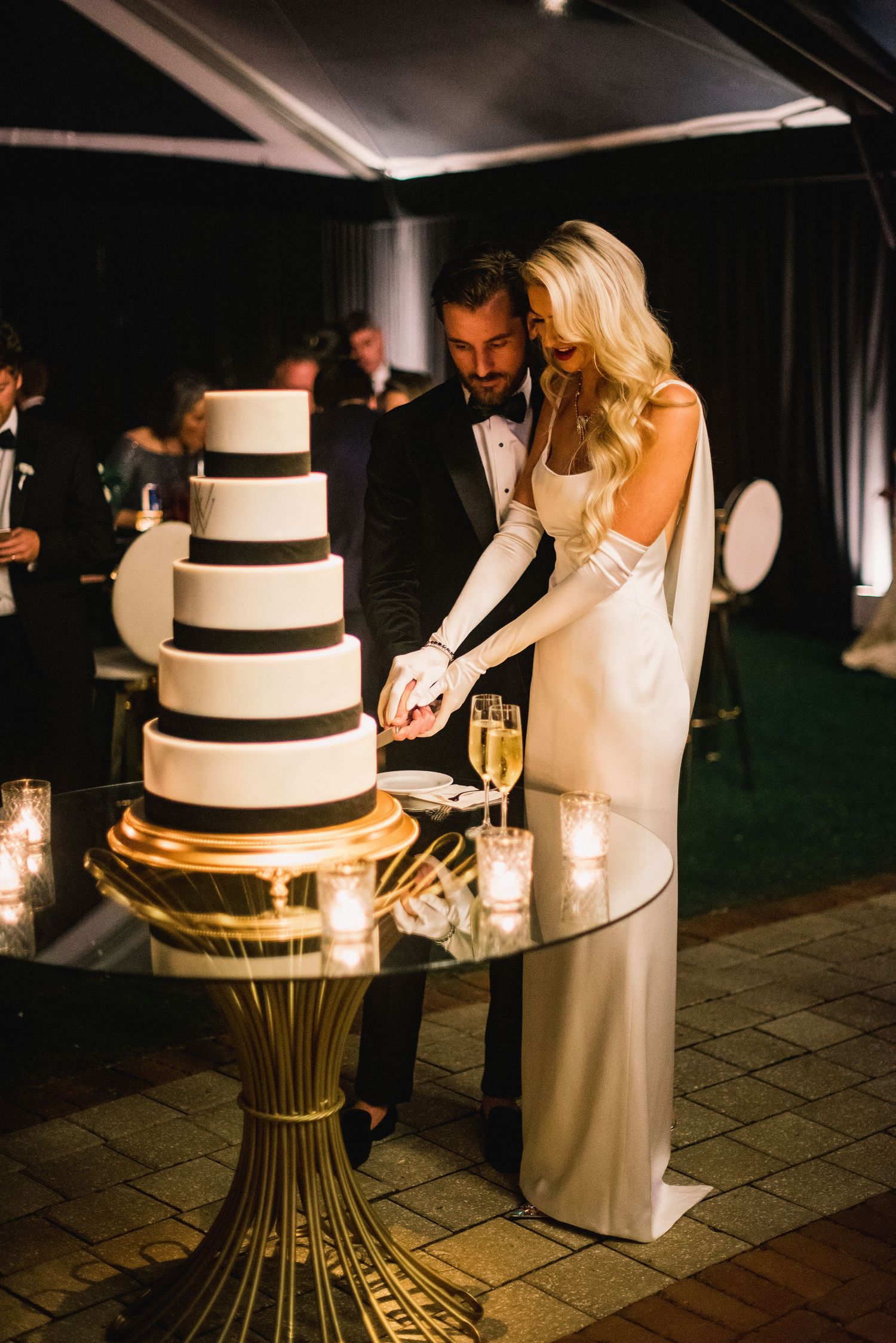 A couple cuts their elegant three-tiered white wedding cake with black ribbons on a gold pedestal stand at night.