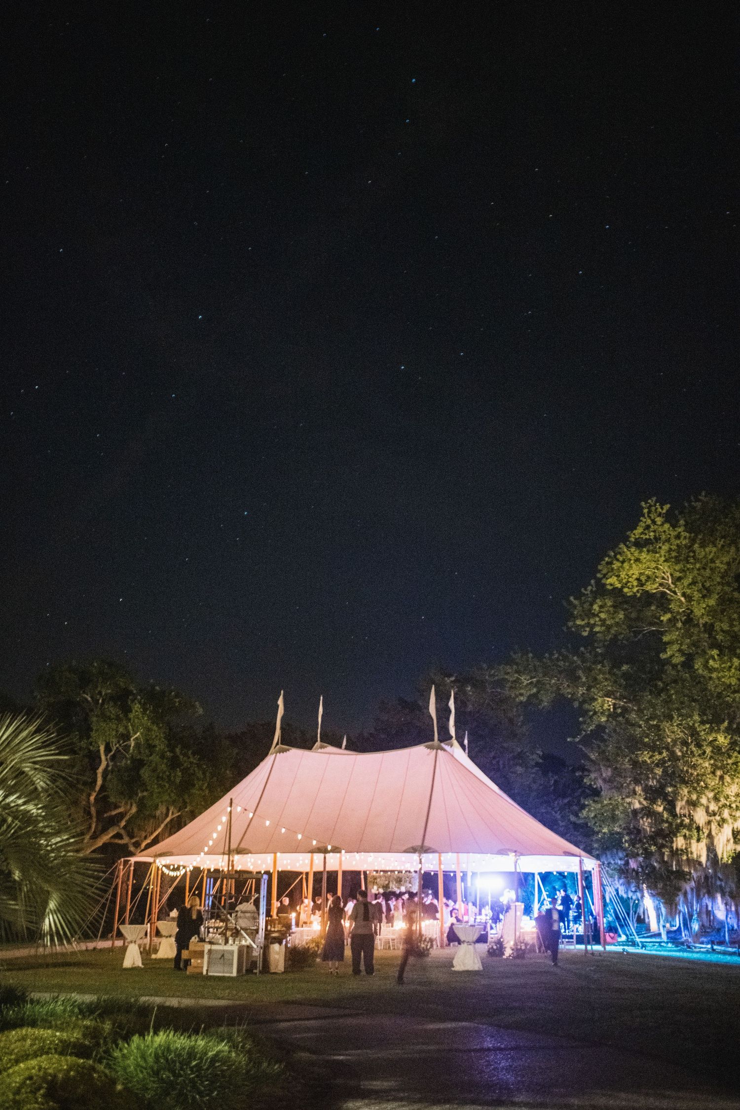 Large white wedding tent illuminated at night with colorful lights and palm trees creating a magical outdoor reception setting.
