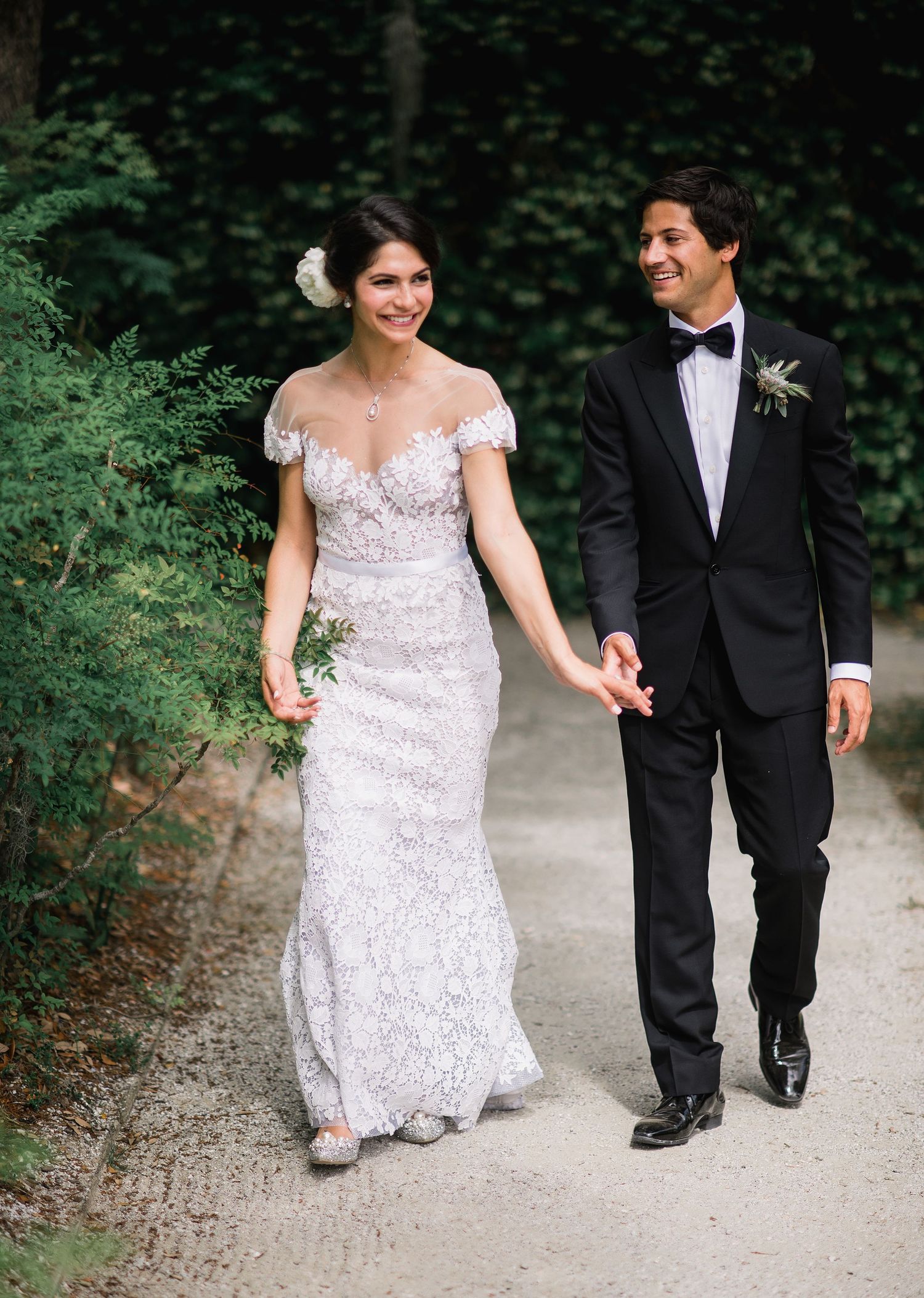 A couple in wedding attire walking together on a garden path, with the bride in a white lace gown and groom in black tuxedo.