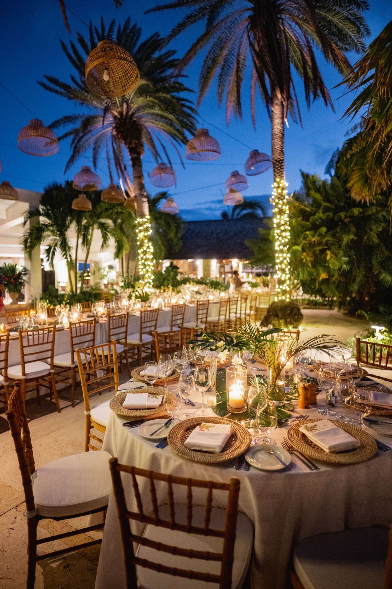 Elegant outdoor wedding reception setup with palm trees and string lights at dusk.