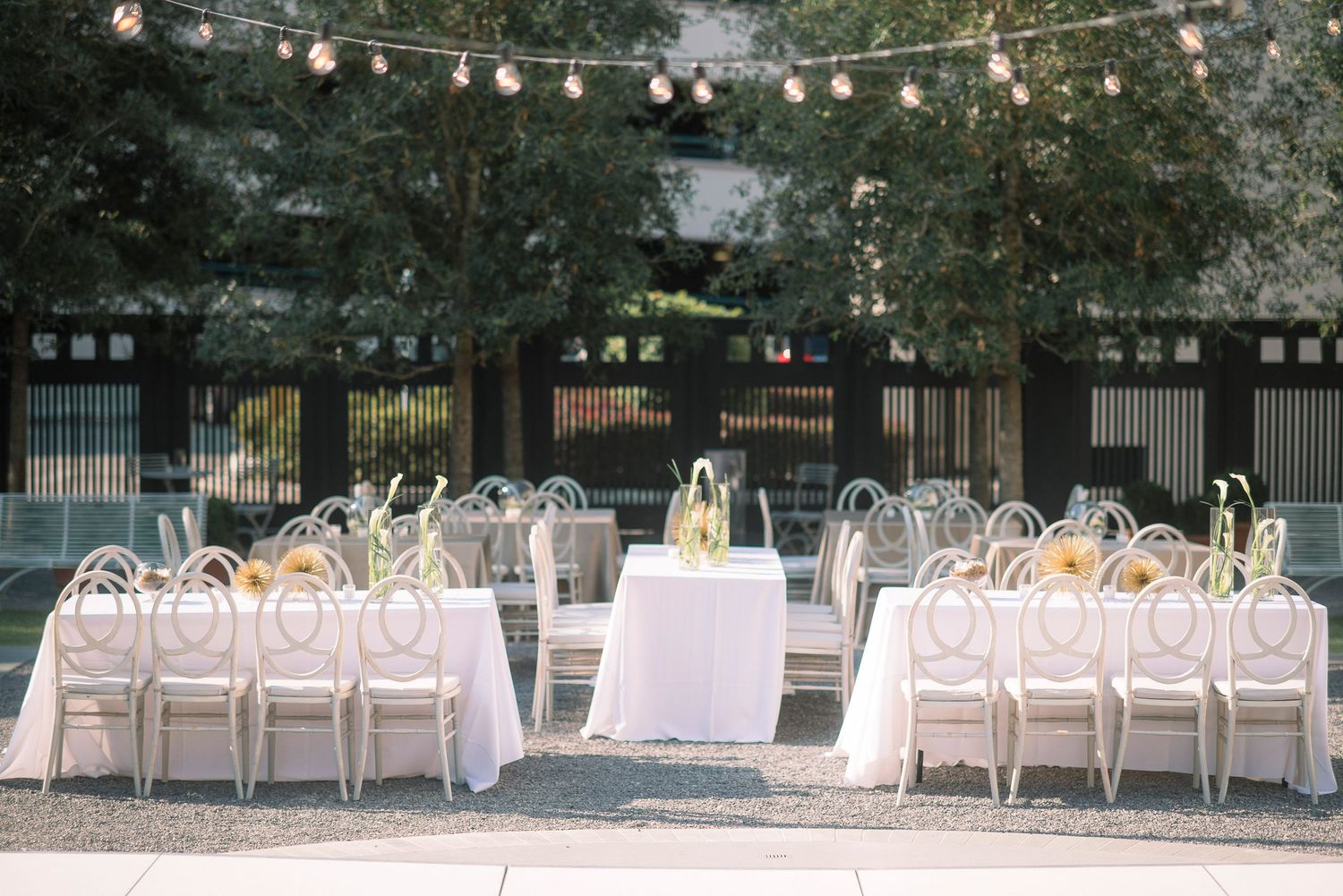 String lights illuminate an outdoor wedding reception setup with white tablecloths and chairs arranged on a patio.