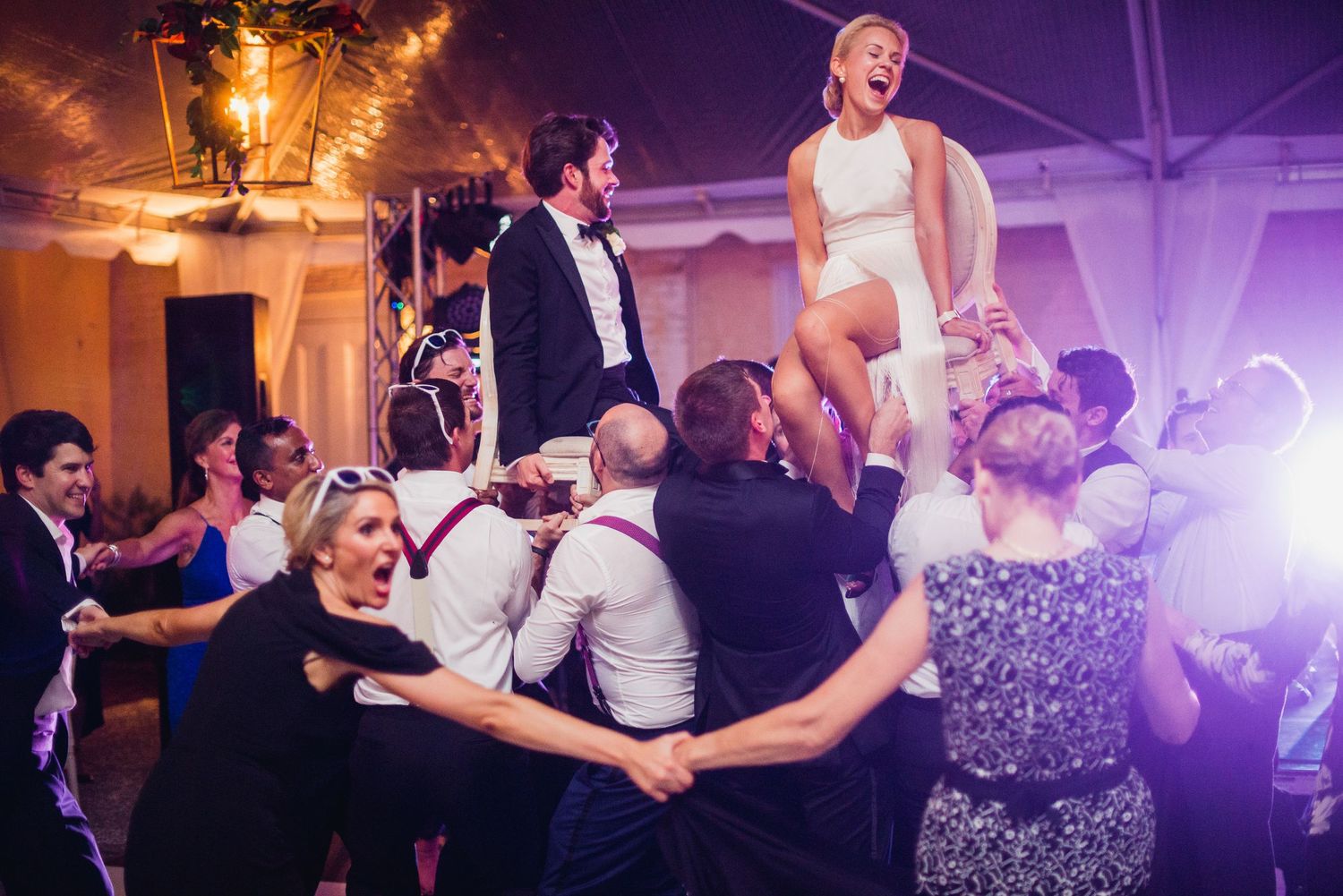 Wedding guests dance and celebrate energetically on a crowded dance floor illuminated by purple and orange lighting.