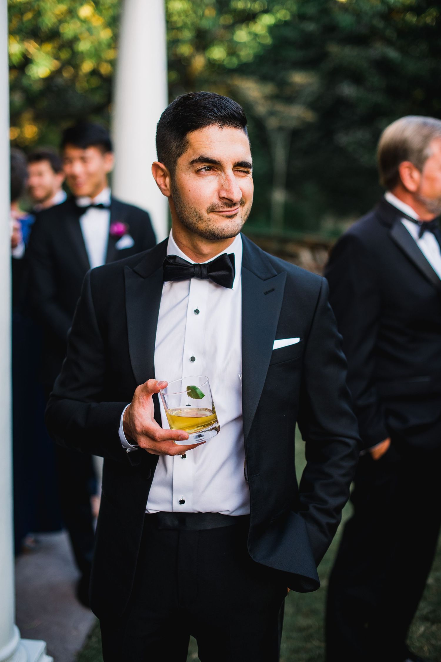 Groomsmen in black tuxedos and bowties gather during an outdoor wedding celebration.