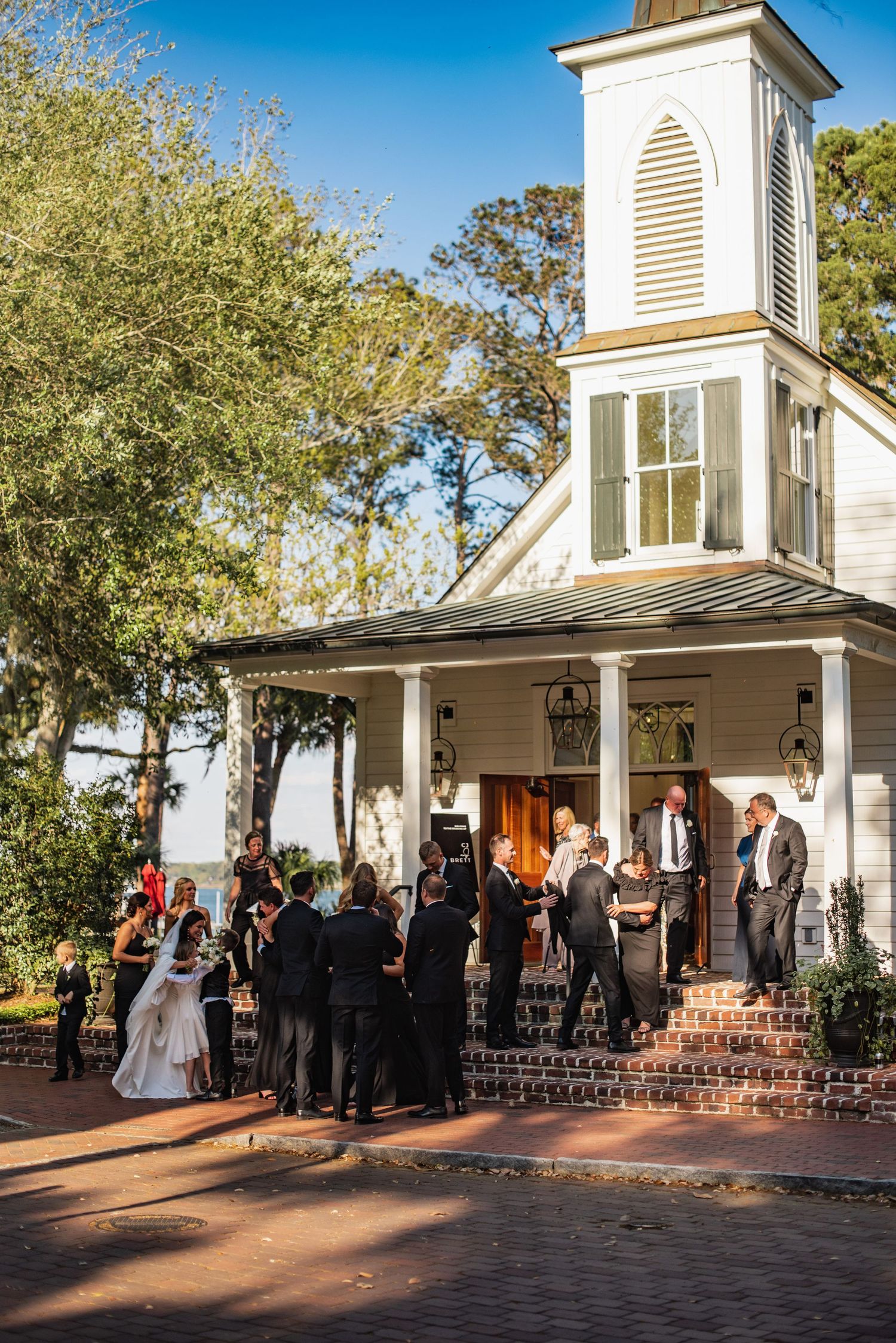 A wedding ceremony procession outside a charming white chapel with brick steps and autumn trees in the background.