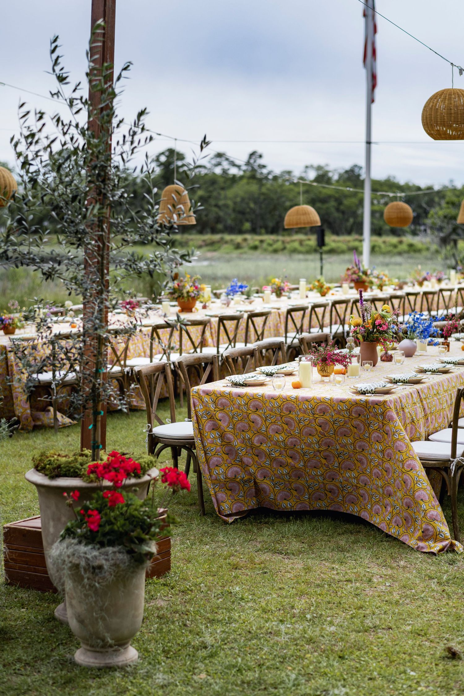 Long rustic dining tables set for an outdoor wedding reception with floral centerpieces and woven pendant lights overhead.