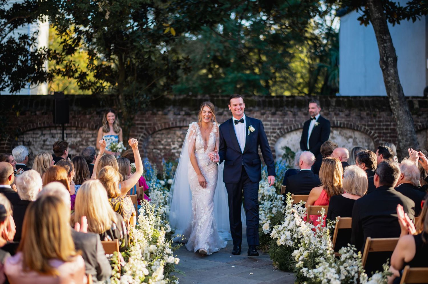 Wedding guests gather outdoors for a ceremony surrounded by lush greenery and white floral arrangements.