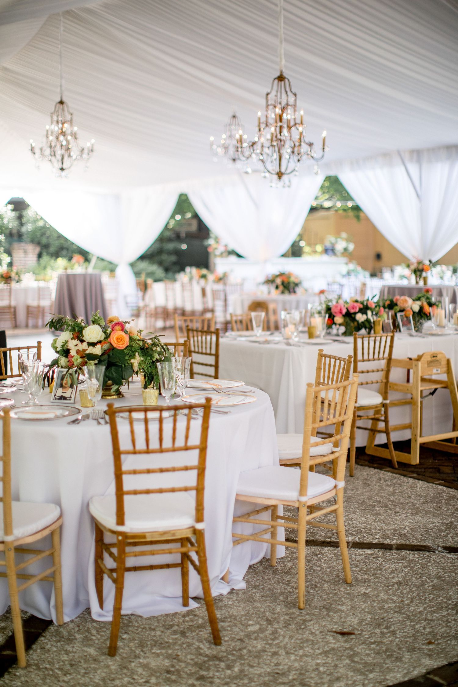 Elegant wedding reception tent with crystal chandeliers, white tablecloths, and gold chiavari chairs arranged around circular tables.