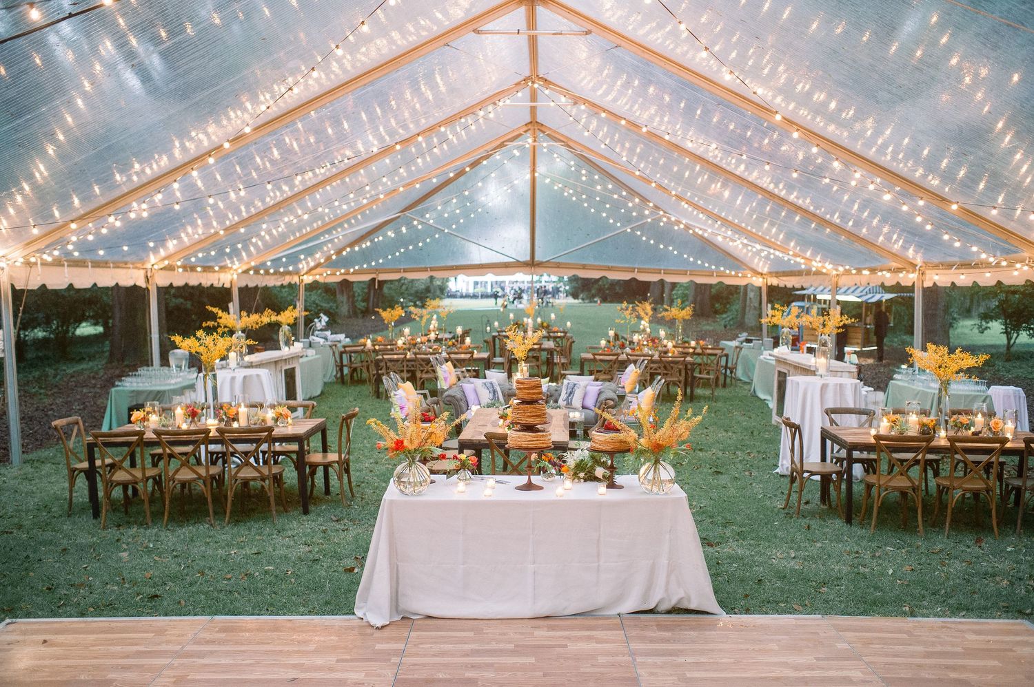 A clear tent wedding reception setup with string lights and wooden tables creating a romantic outdoor atmosphere.
