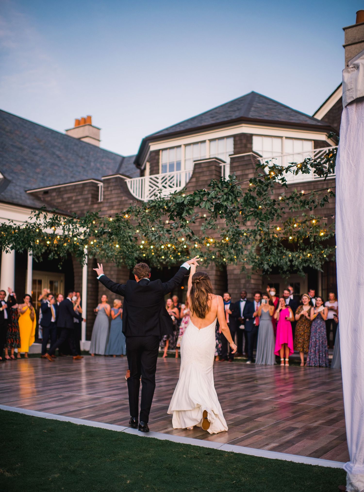 A couple celebrates on an outdoor dance floor at dusk with string lights overhead and wedding guests watching from the sides.
