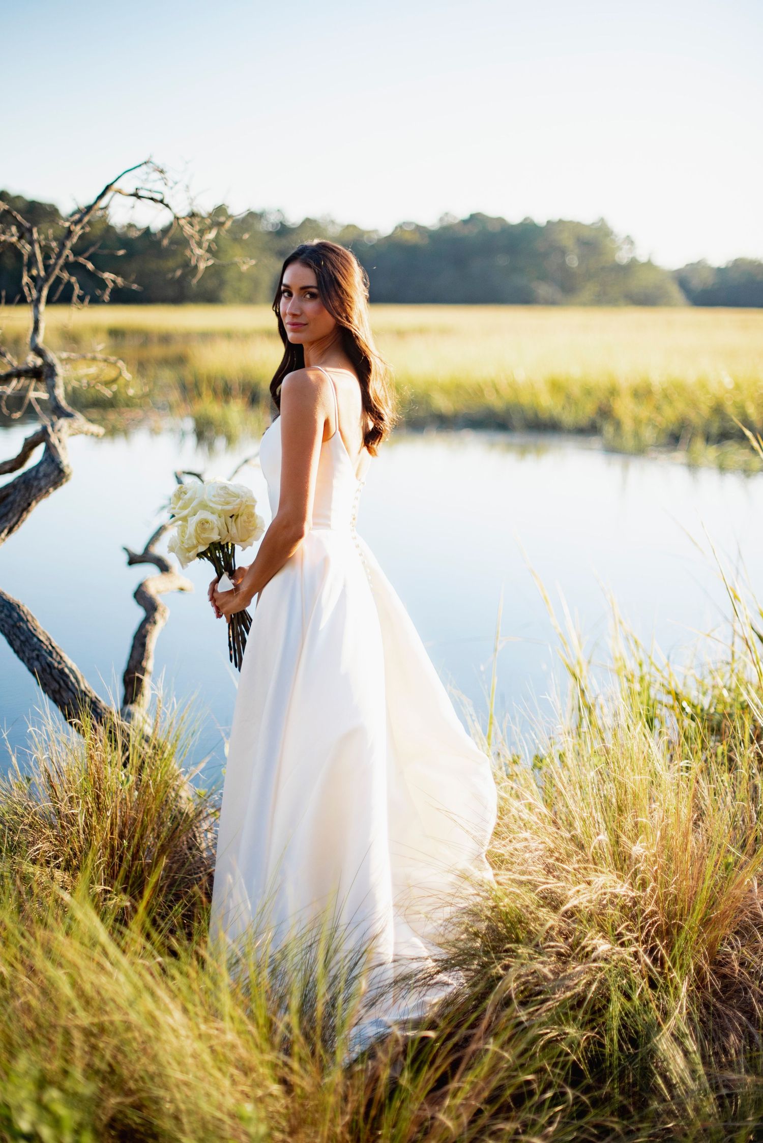 A flowing white wedding dress catches sunlight as it moves against tall marsh grasses beside a tranquil lake.