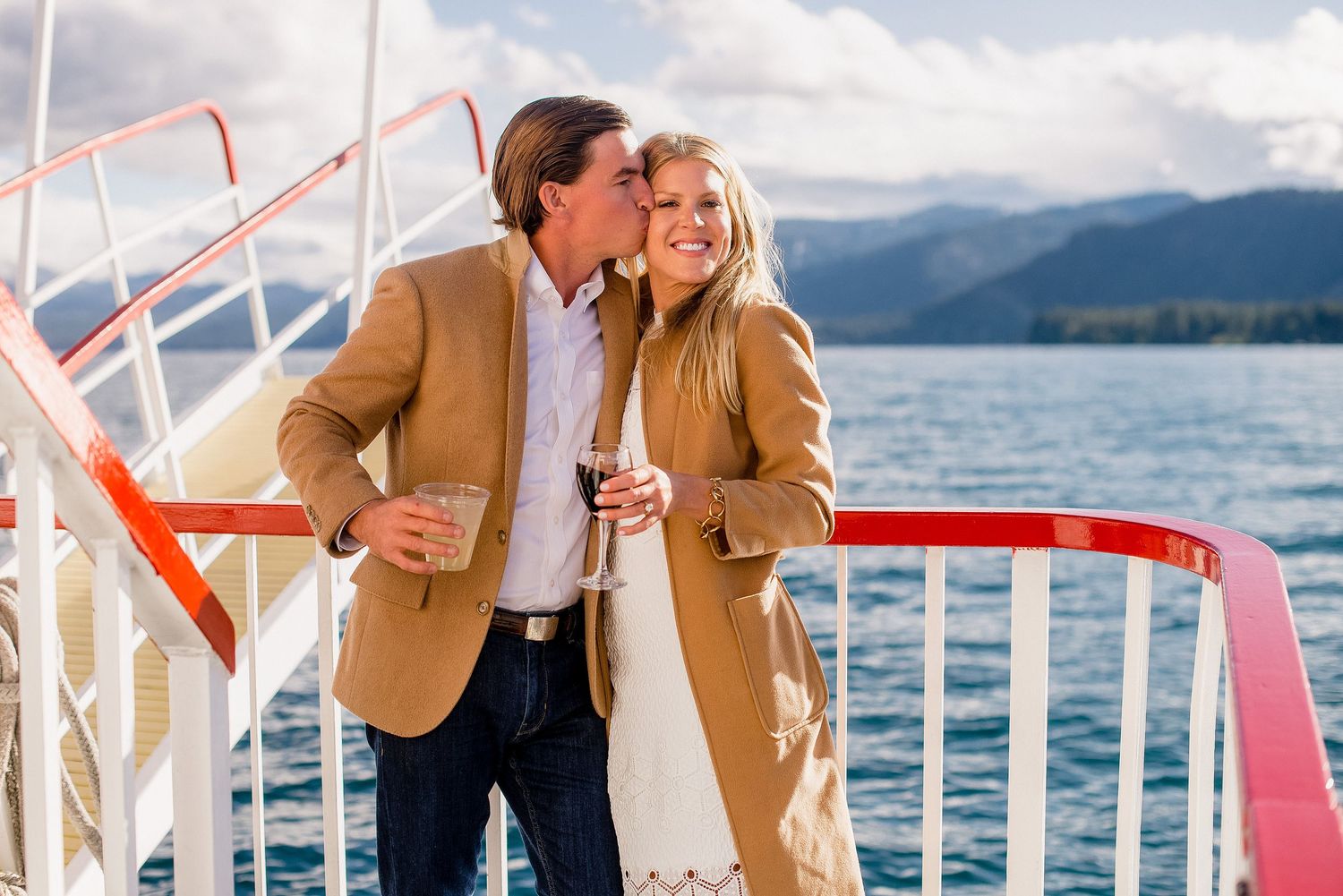 Couple in tan jackets sharing drinks while enjoying scenic mountain lake views from boat deck railing.