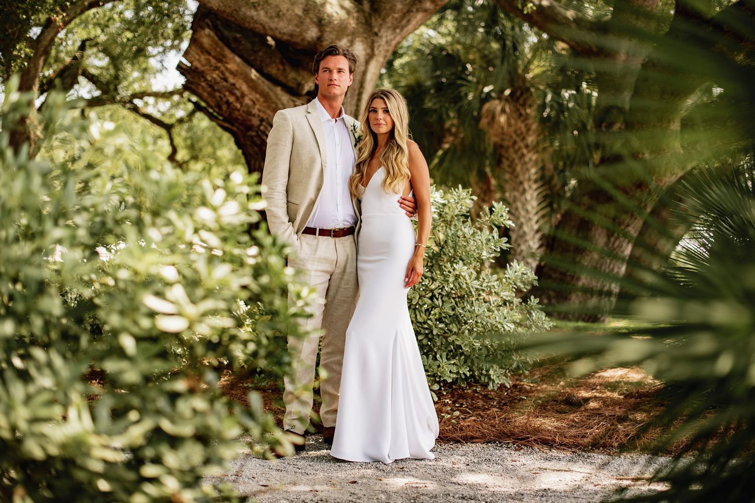 A couple in white outfits walks hand in hand through a lush garden path on a sunny afternoon.