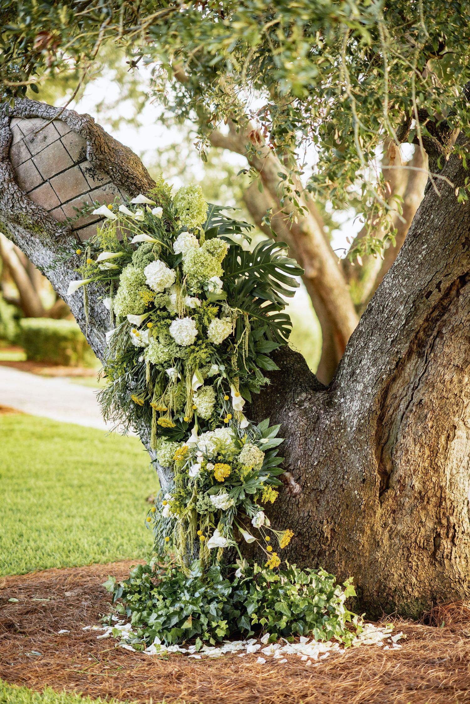 Beautiful floral wedding arrangement with white and yellow flowers cascading down a rustic tree trunk in an outdoor garden setting.