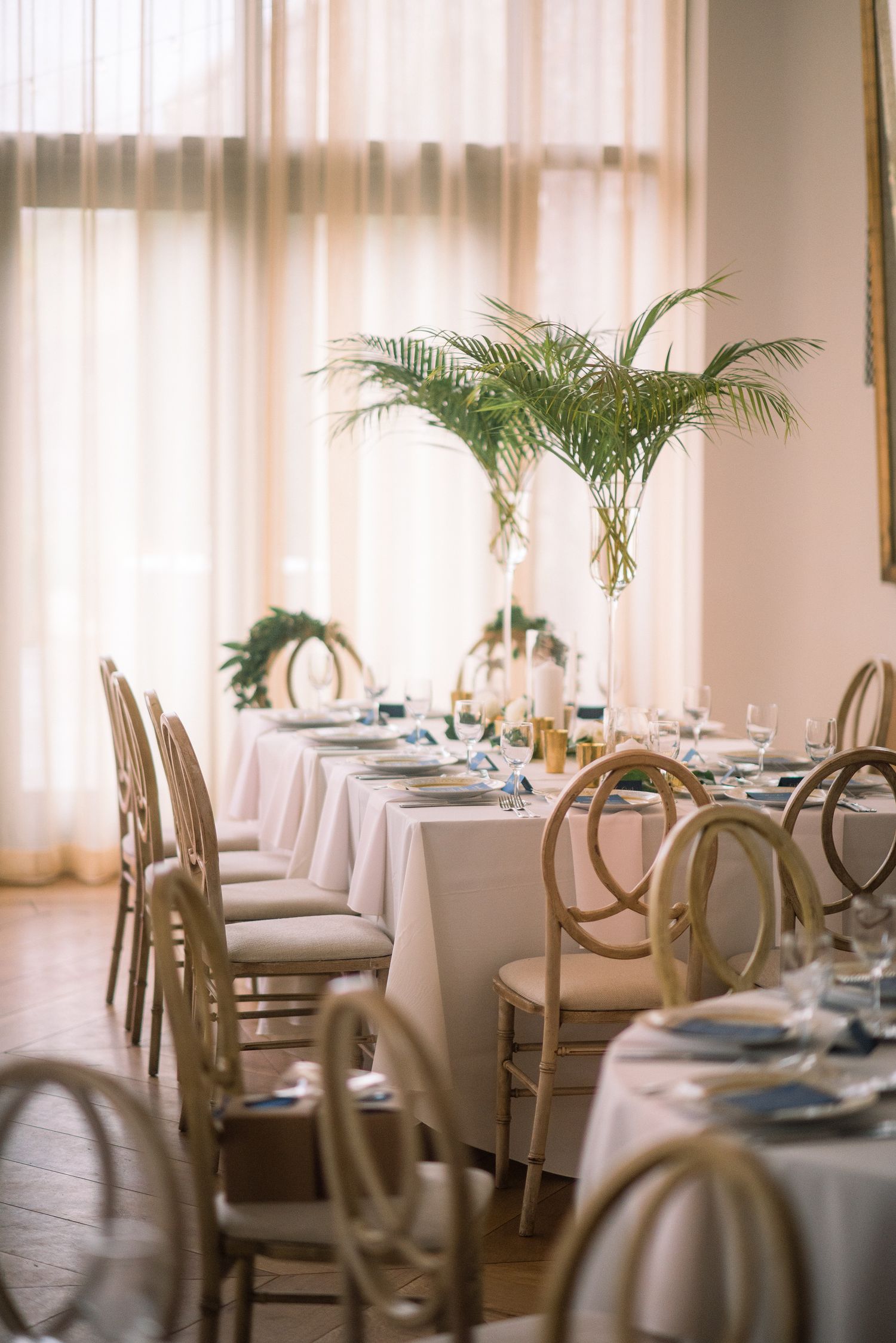 Elegant wedding reception tables set with white linens, palm fronds and round back chairs in a bright sunlit venue.