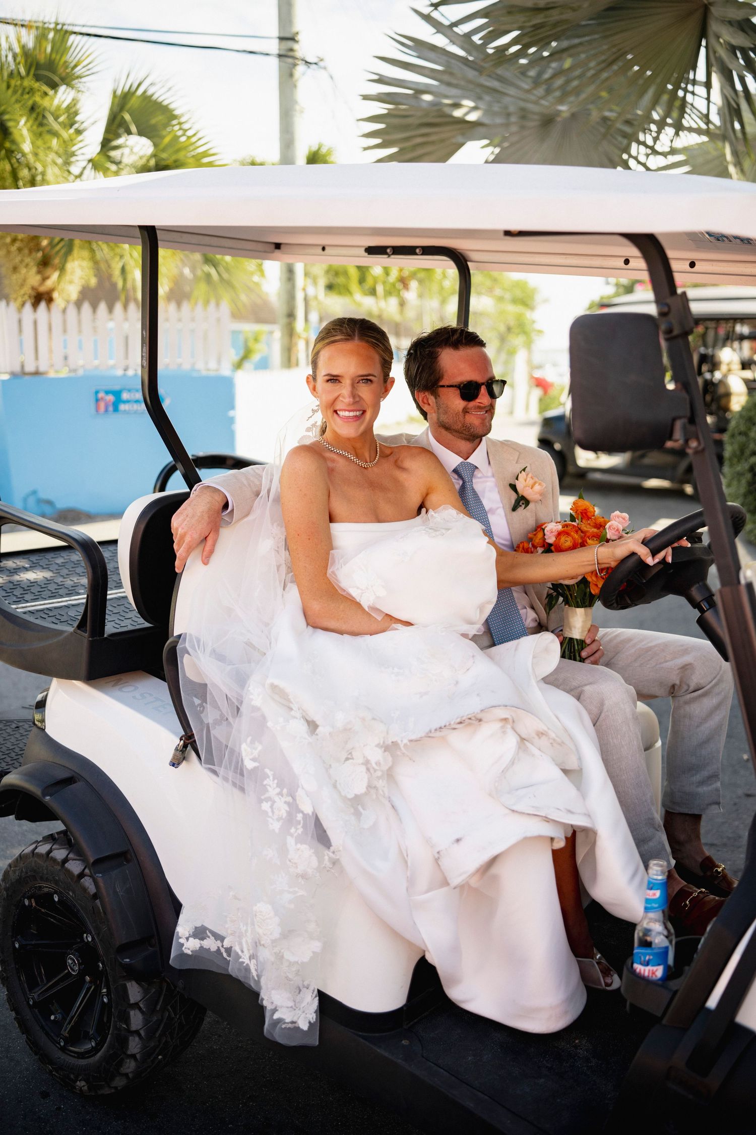 A wedding couple rides in a golf cart decorated with white fabric and orange flowers.