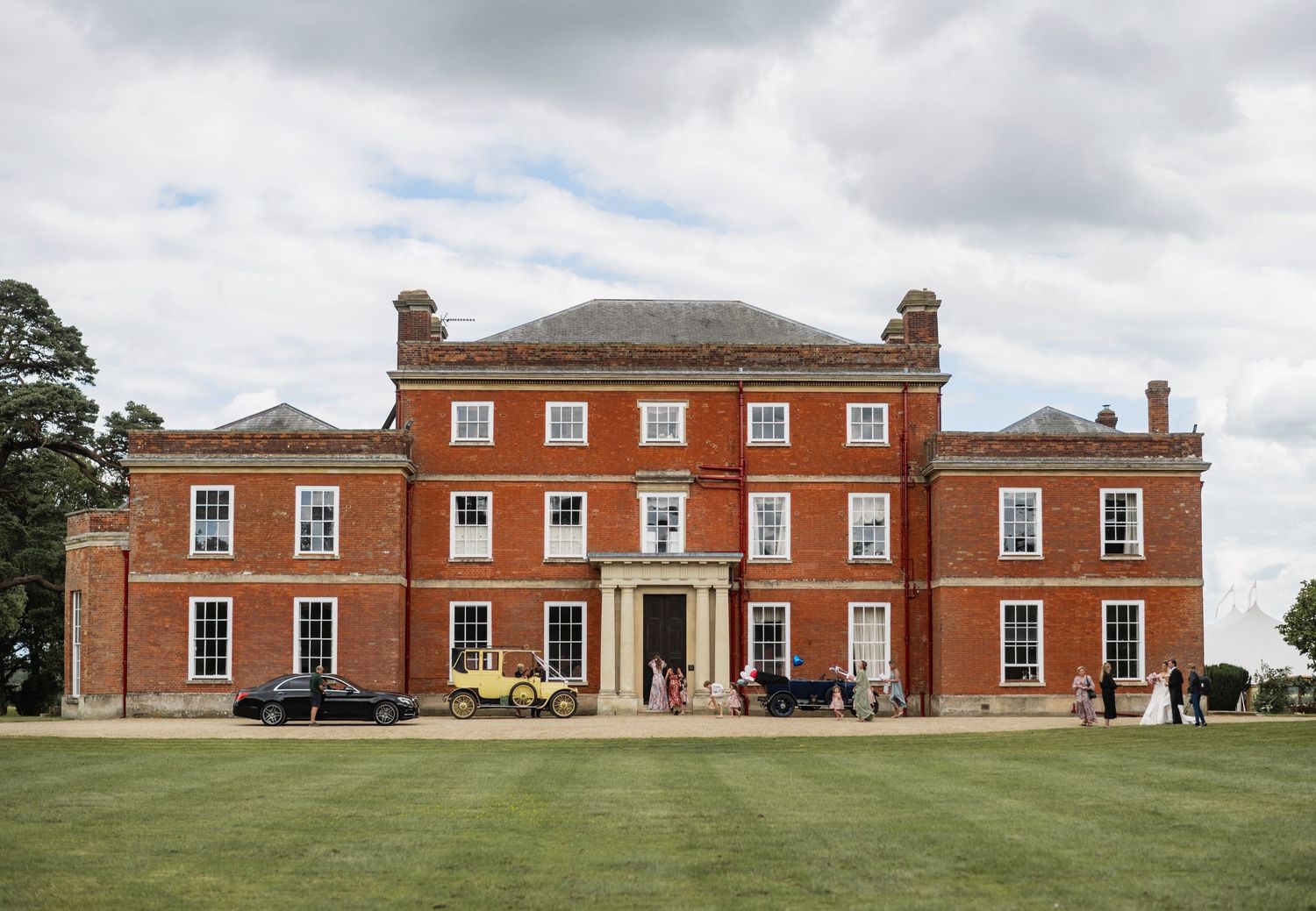 Grand red brick Georgian manor house with symmetrical architecture set on manicured lawn under cloudy sky. Cambridgeshire, EN
