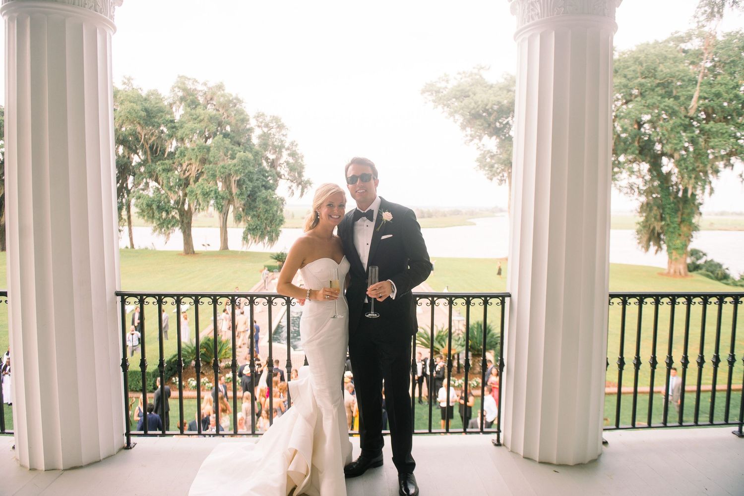 Elegant wedding couple shares a romantic moment between white pillars on an outdoor veranda overlooking a scenic landscape.