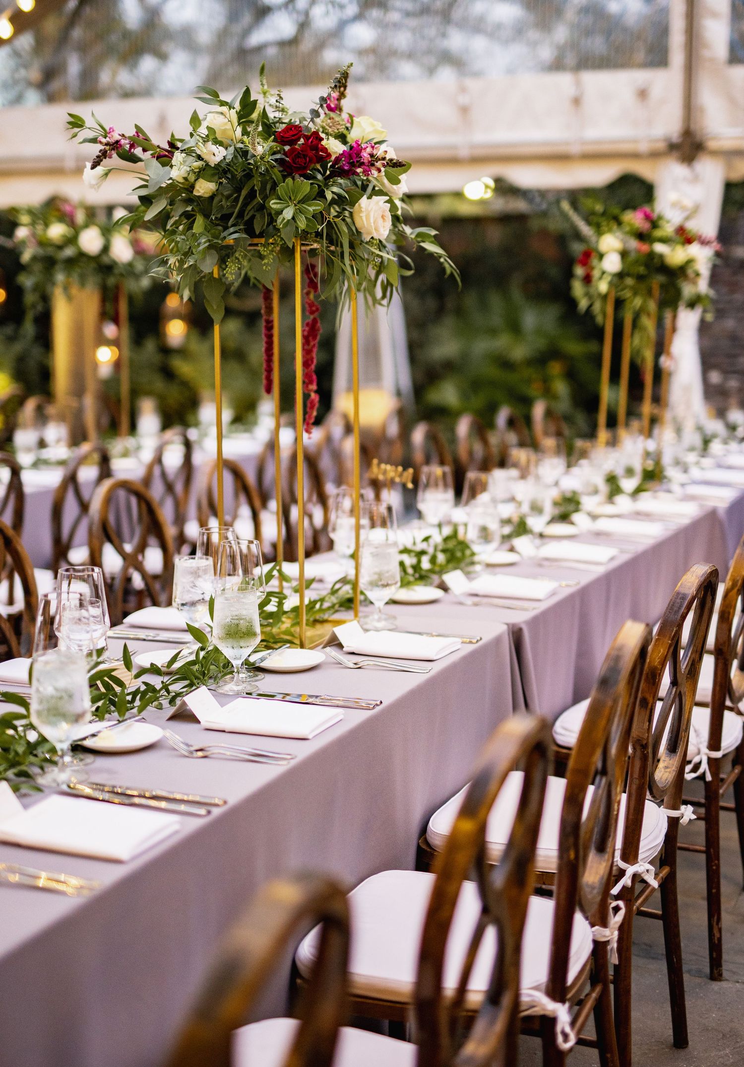 Elegant wedding reception table with tall floral centerpieces, wooden cross-back chairs, and a lavender tablecloth.