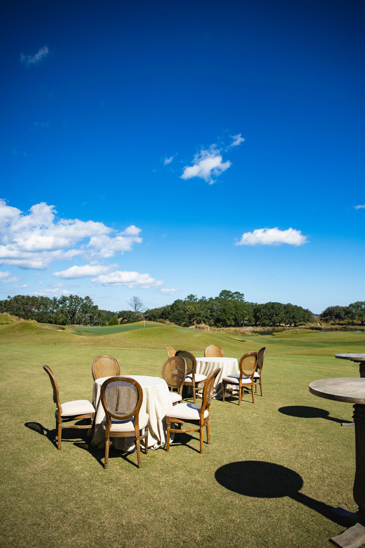 Wooden patio chairs and table set arranged on a lush green golf course under bright blue sky with white clouds.