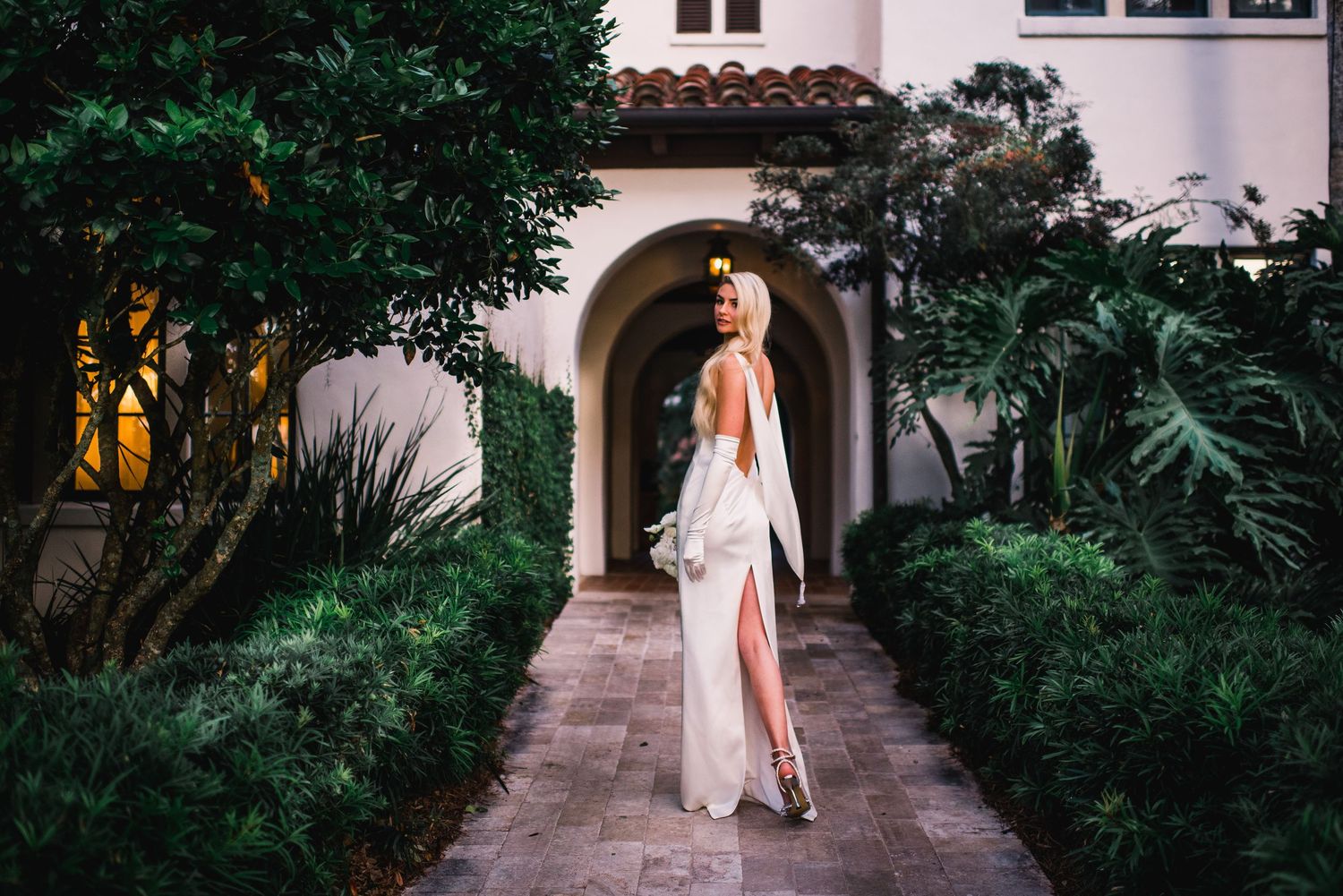 A figure in a flowing white dress walks along a lush garden path lined with tropical greenery and stone archways.