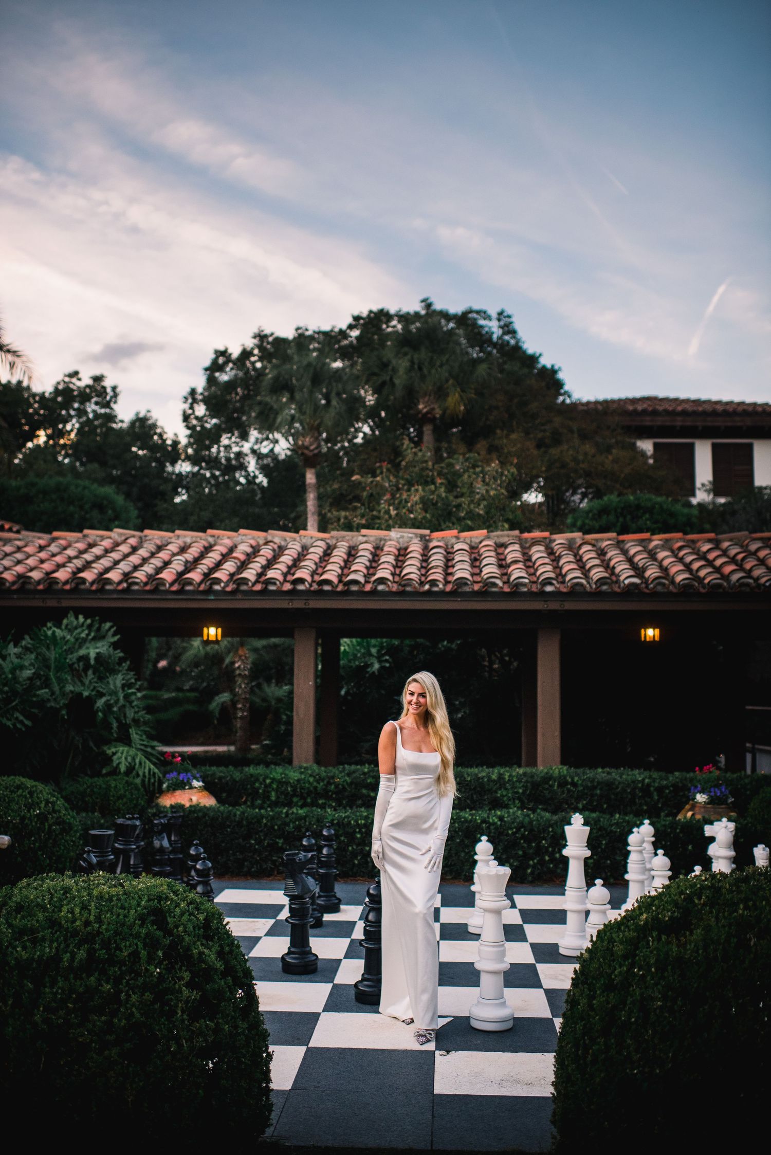 Bride stands on outdoor chess board surrounded by topiary gardens at dusk.