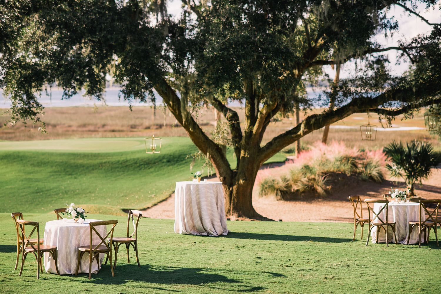 Outdoor wedding reception setup with wooden chairs and white tablecloths under a tree in a rustic garden setting.