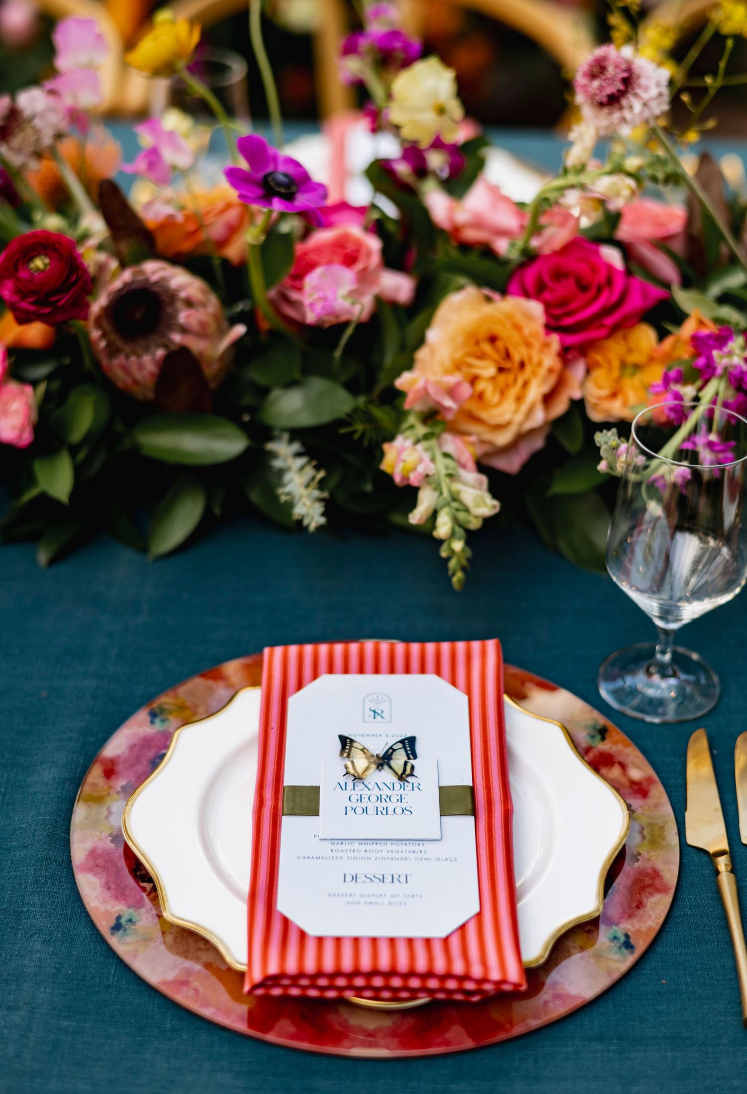 Elegant place settings with red and white striped napkins and colorful floral arrangements on dark teal tablecloth.