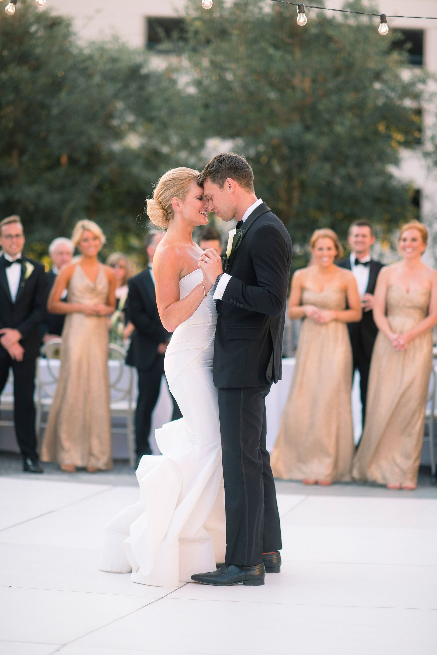 A couple shares their first dance at an outdoor wedding reception with bridesmaids in champagne dresses looking on.