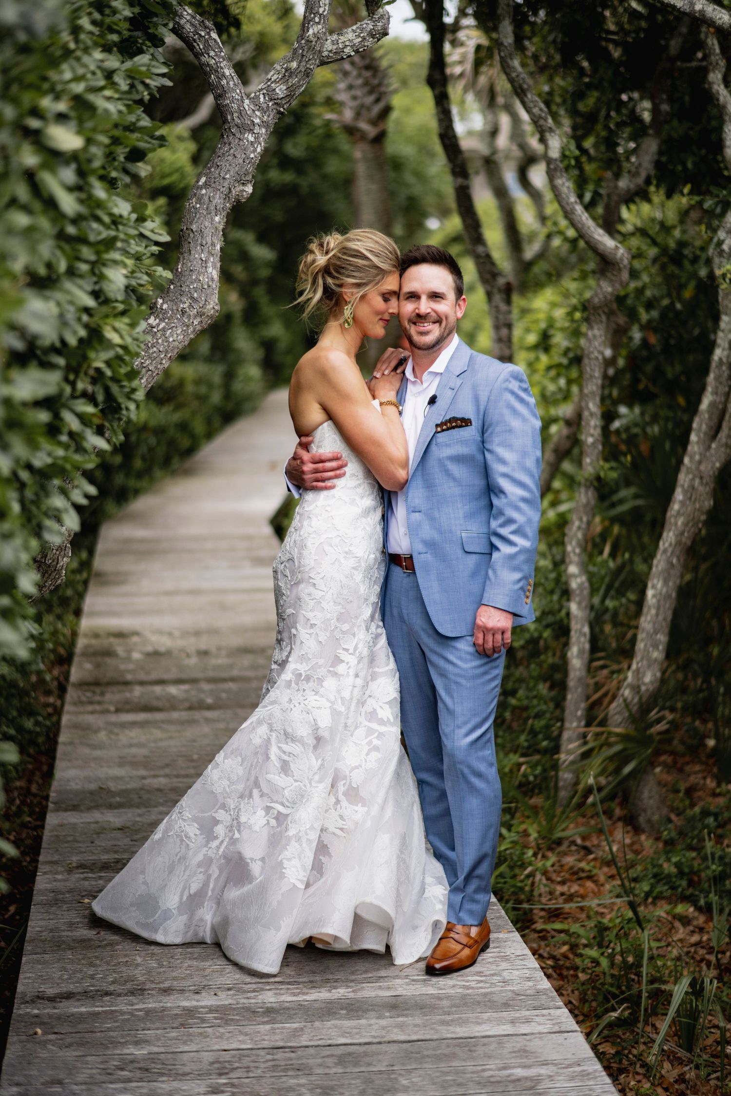 A couple in wedding attire share an intimate moment on a wooden boardwalk surrounded by lush greenery.