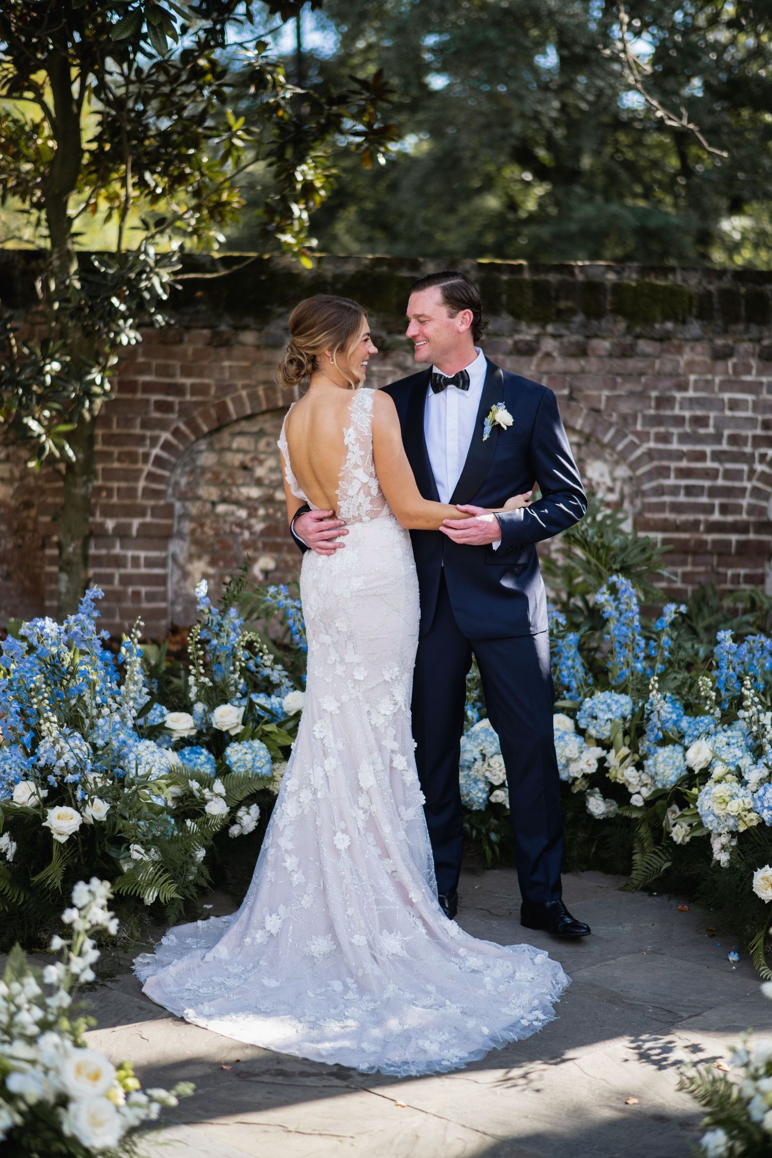 A romantic wedding couple embraces in a garden setting with blue and white flowers against a brick wall.