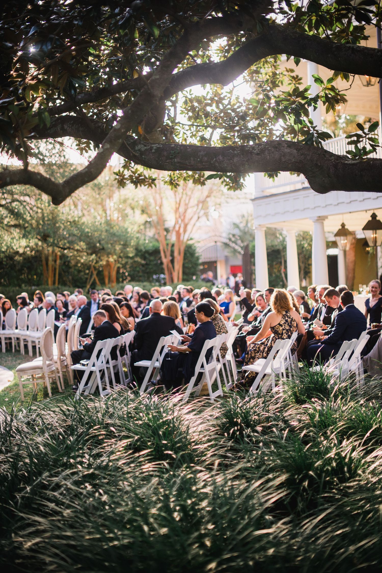 Outdoor wedding ceremony setup with rows of white chairs surrounded by lush greenery and trees at sunset.