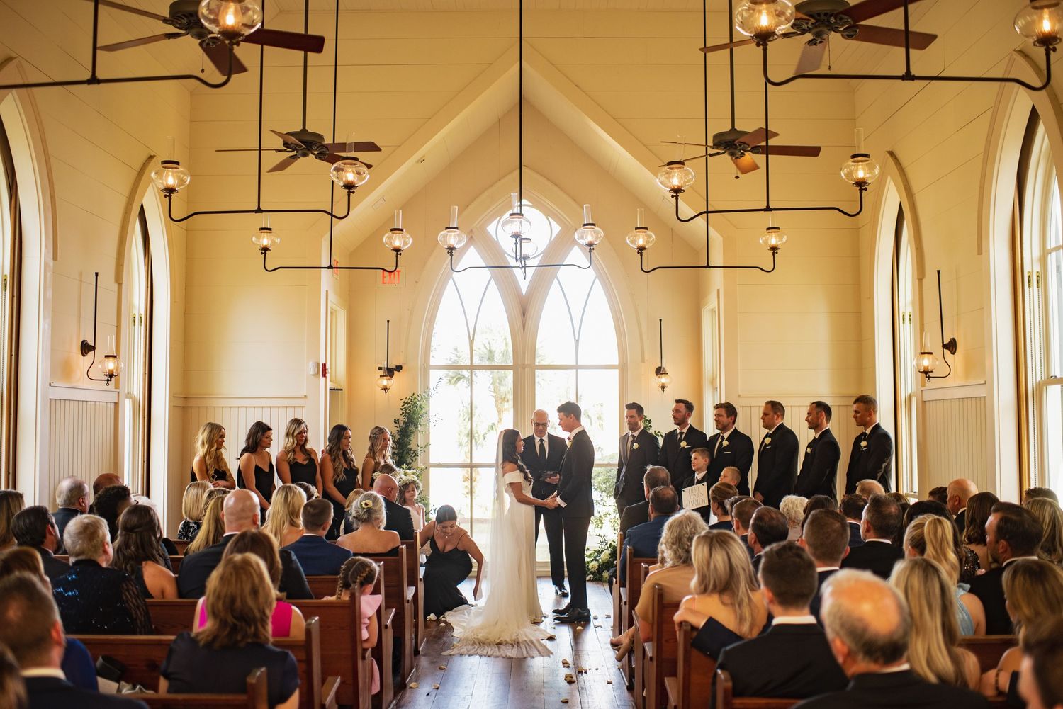 A wedding ceremony takes place in a bright church with arched windows and multiple ceiling fans.