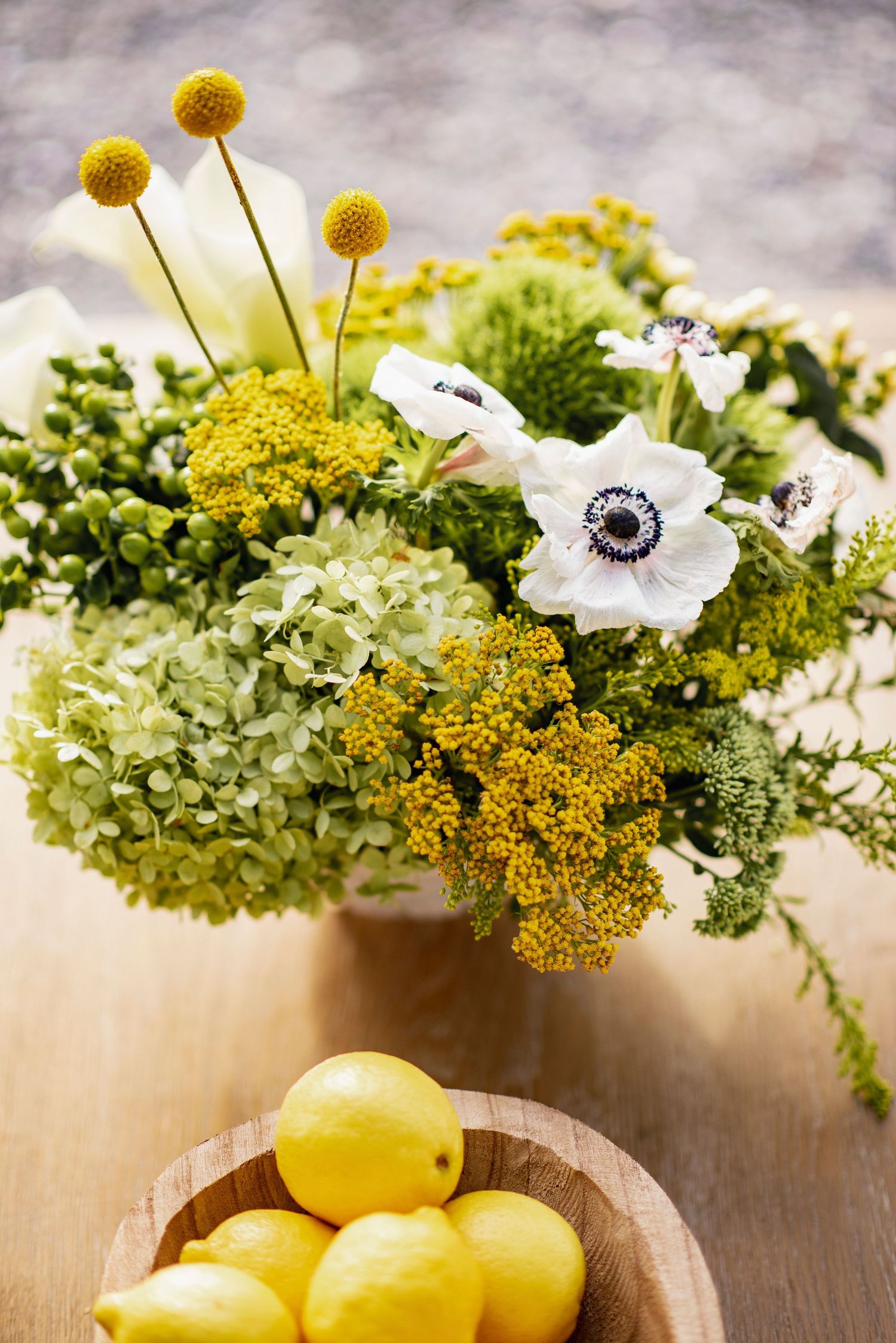 Fresh yellow lemons and white anemone flowers with yellow yarrow in rustic floral arrangement on wooden table.