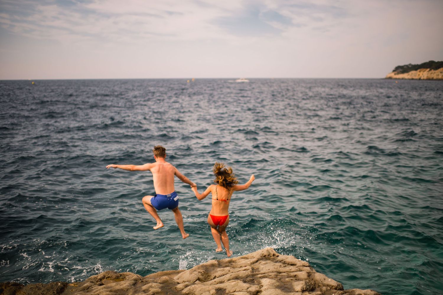 People jumping off a rocky cliff into turquoise Mediterranean waters at sunset.