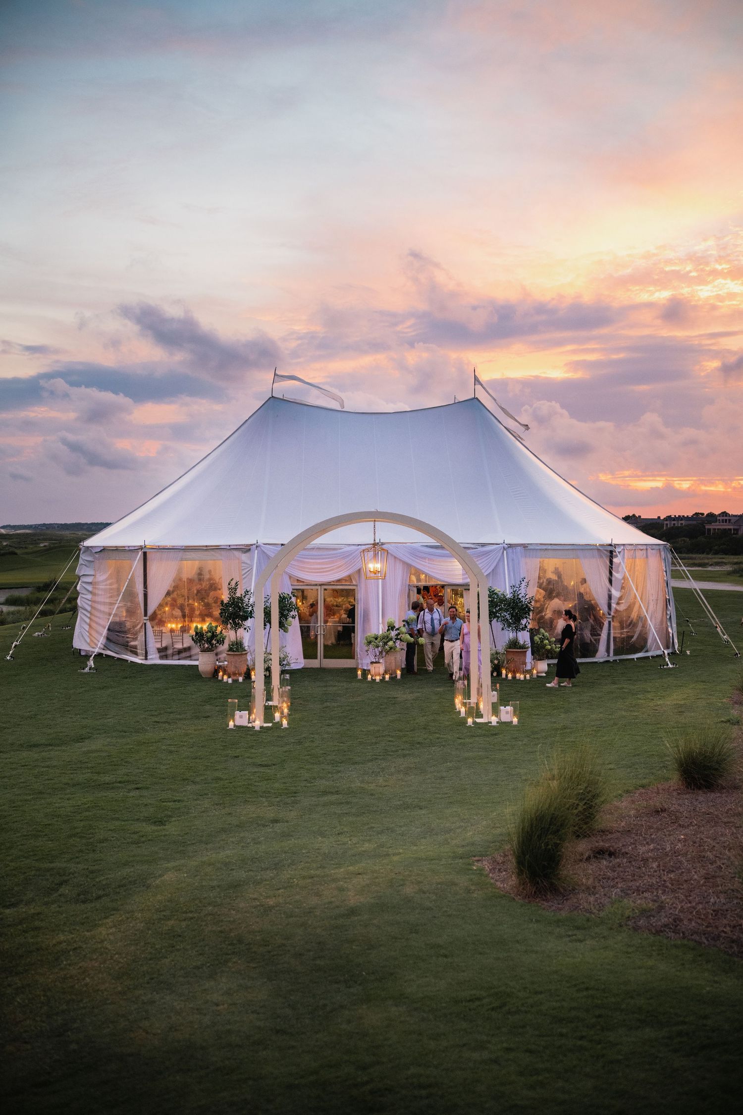 White wedding tent illuminated at sunset with glowing lights and decorative entrance on a grassy field.