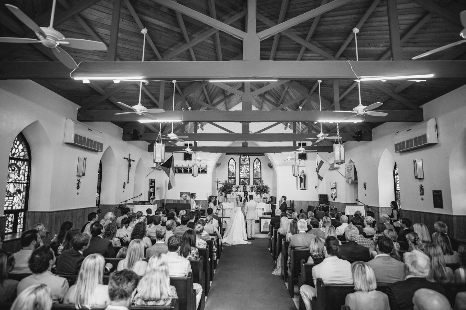 Black and white photo of a church wedding ceremony with guests seated in wooden pews under vaulted ceilings.