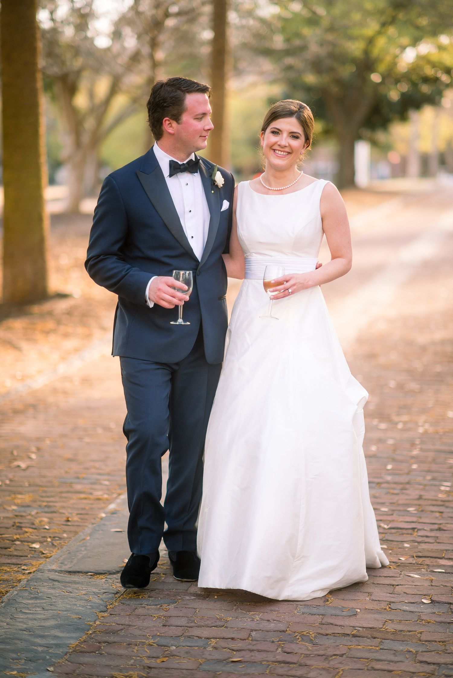 Couple in wedding attire walking together on a cobblestone path during golden hour in an autumn setting.