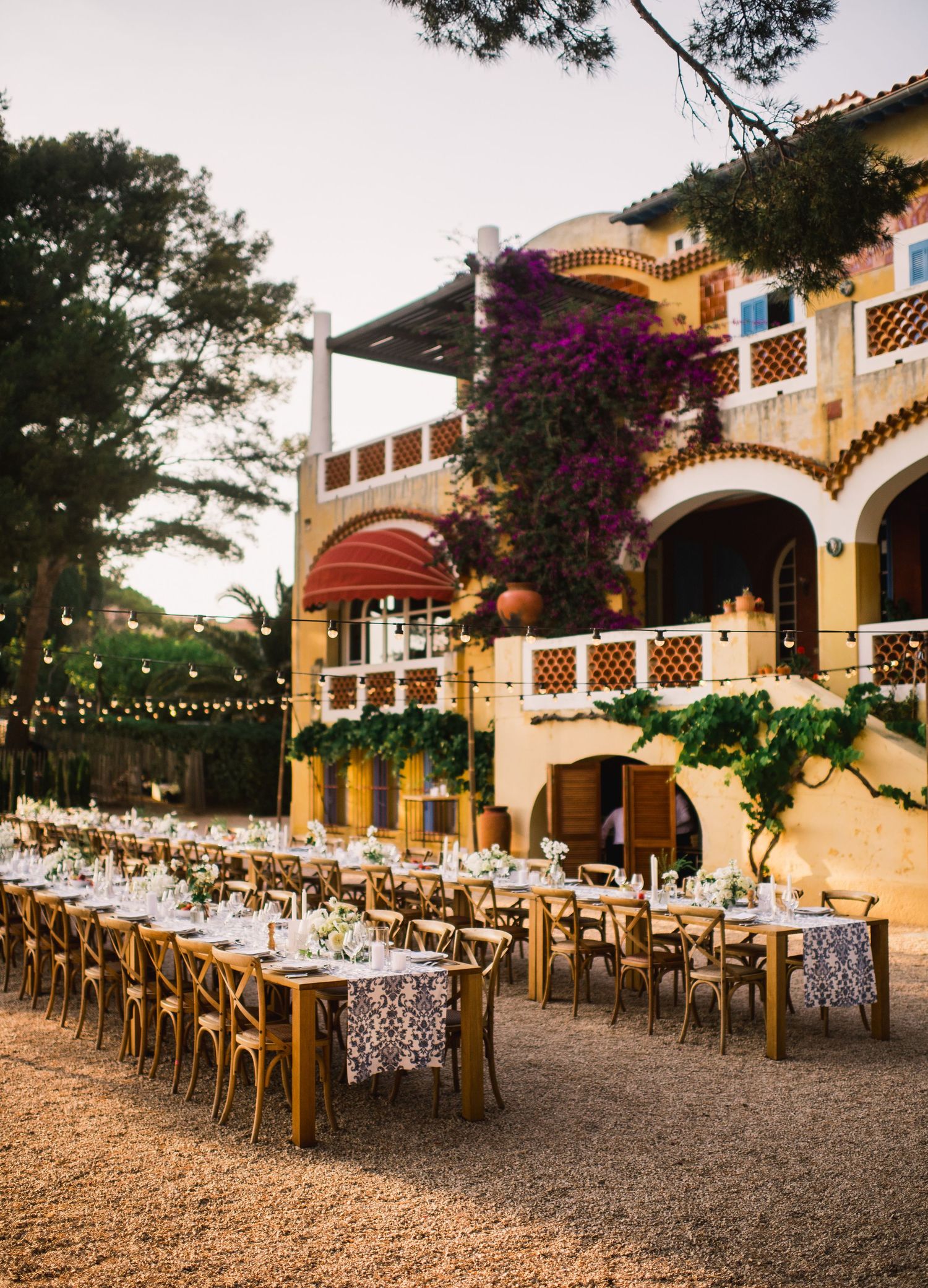 Elegant outdoor wedding reception setup with long tables, floral tablecloths, and string lights under Spanish-style arches.