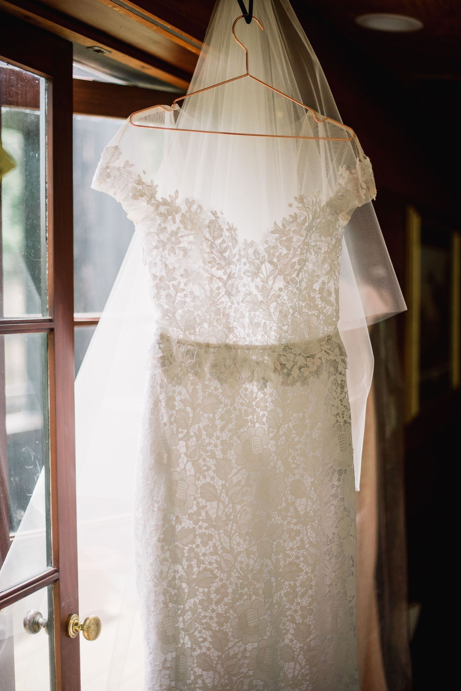 An elegant white lace wedding dress with off-shoulder detail hangs by a window in natural light.