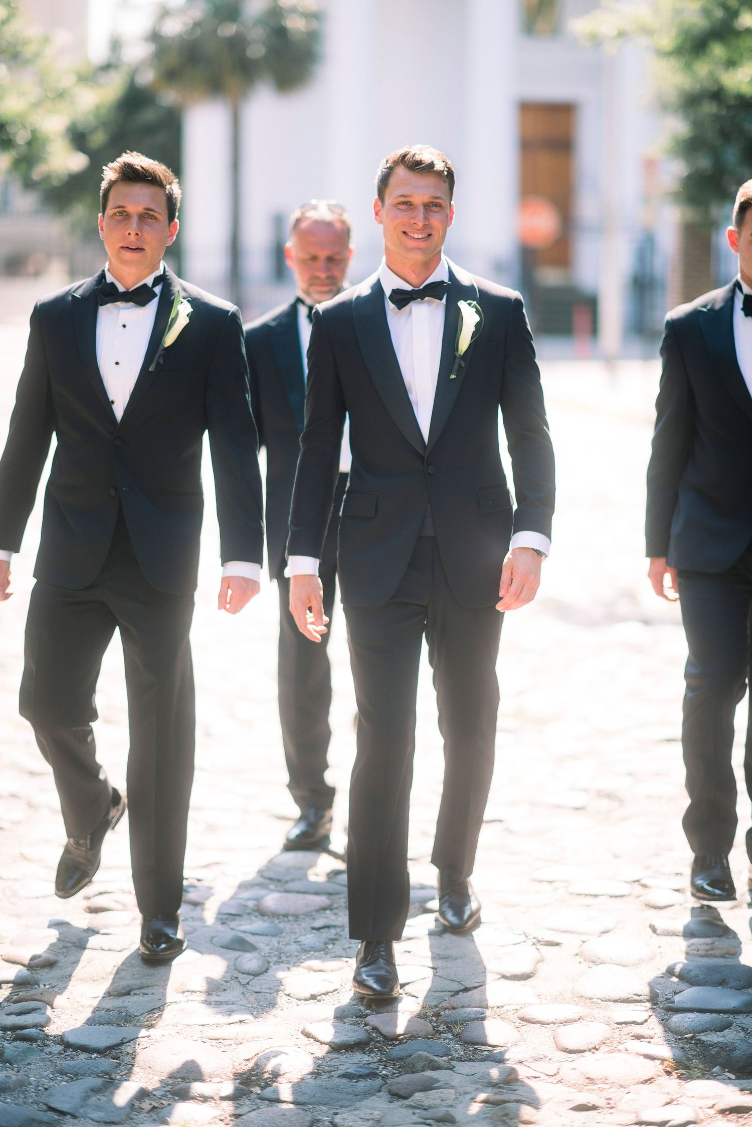 Creative wedding photo showing groomsmen's legs and shoes walking in a line on cobblestones in black tuxedos.