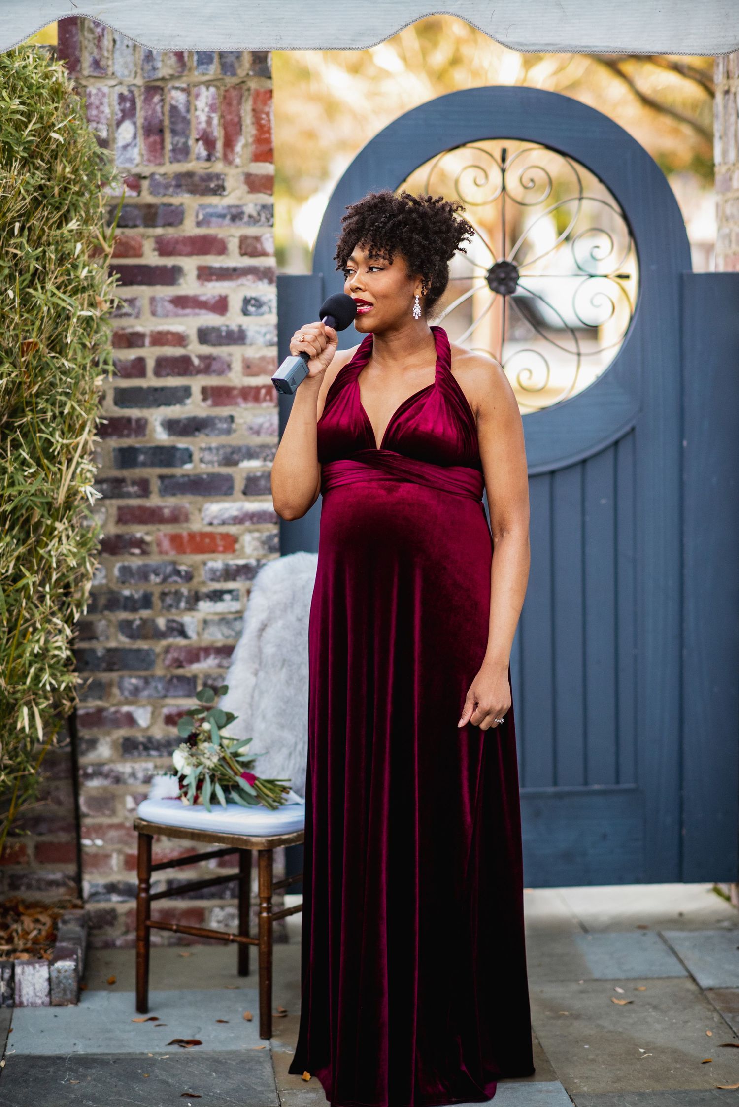 A person in a burgundy velvet dress poses against a brick wall and blue door in an outdoor setting.