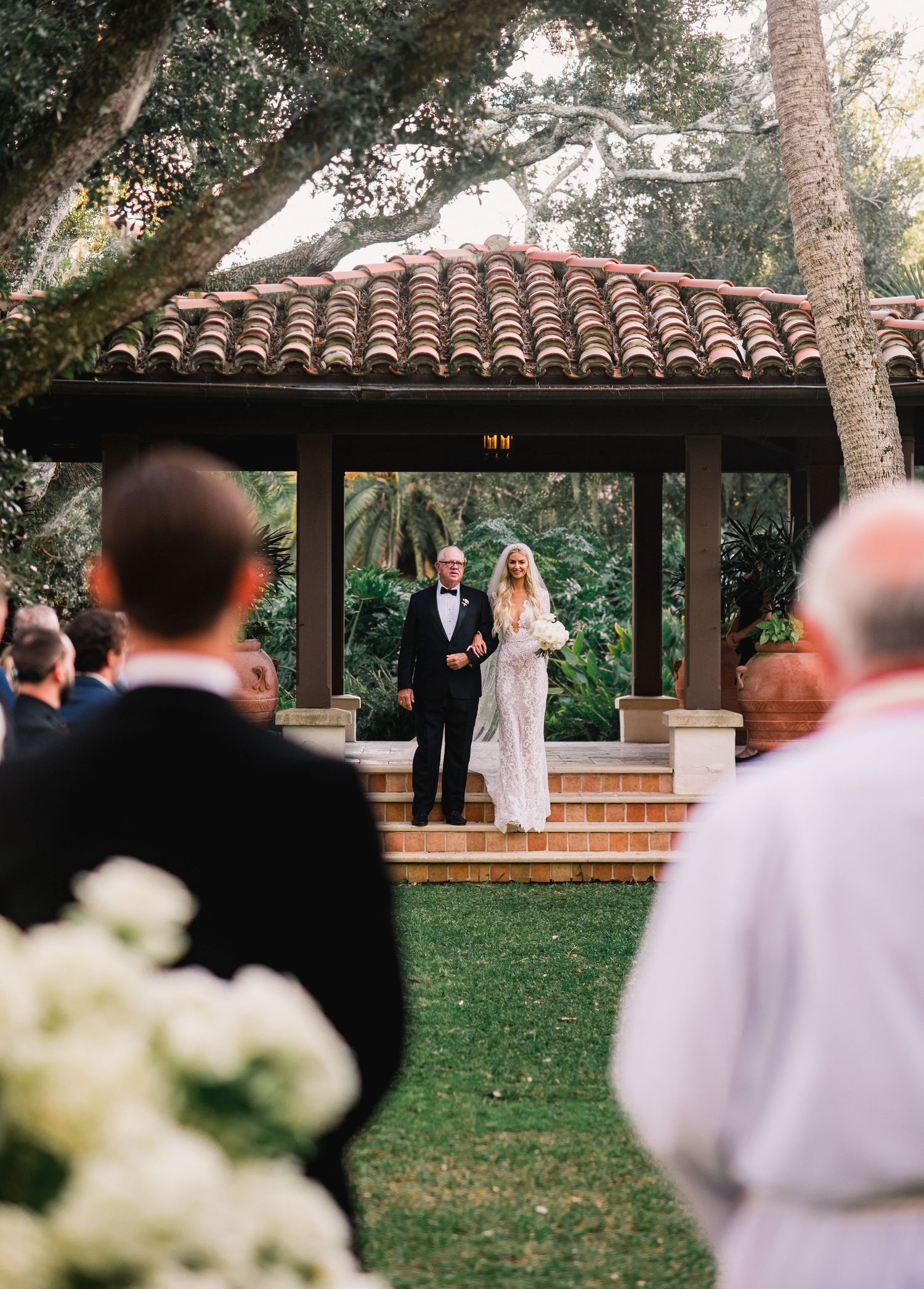 An outdoor wedding ceremony takes place under a wooden pergola with Spanish tile roofing on a grassy lawn.