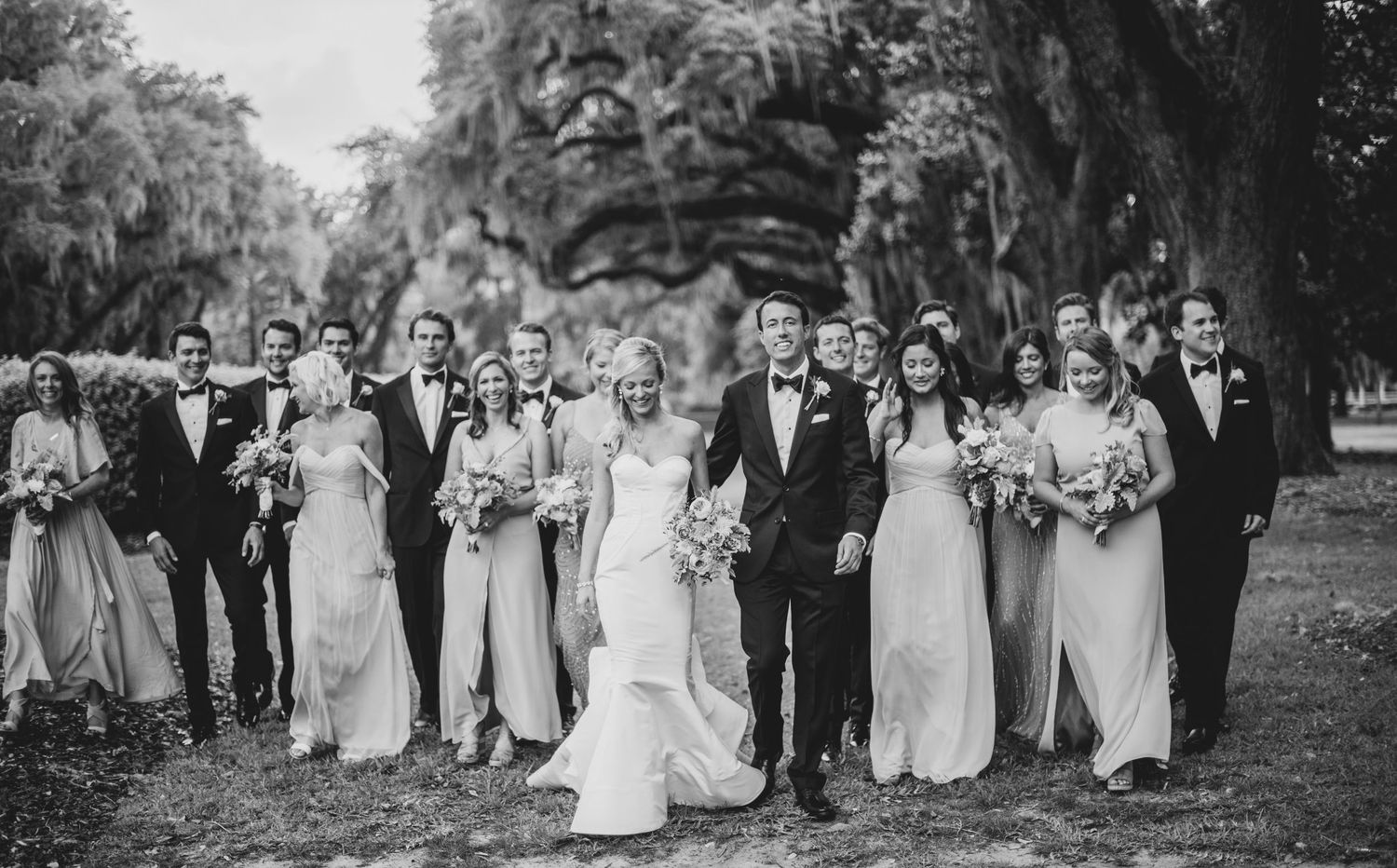 Black and white wedding party poses together outdoors in a scenic garden setting with trees in the background.
