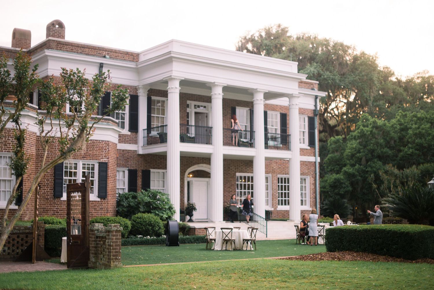 A grand white colonial style building with columns and portico stands on a manicured lawn surrounded by trees.