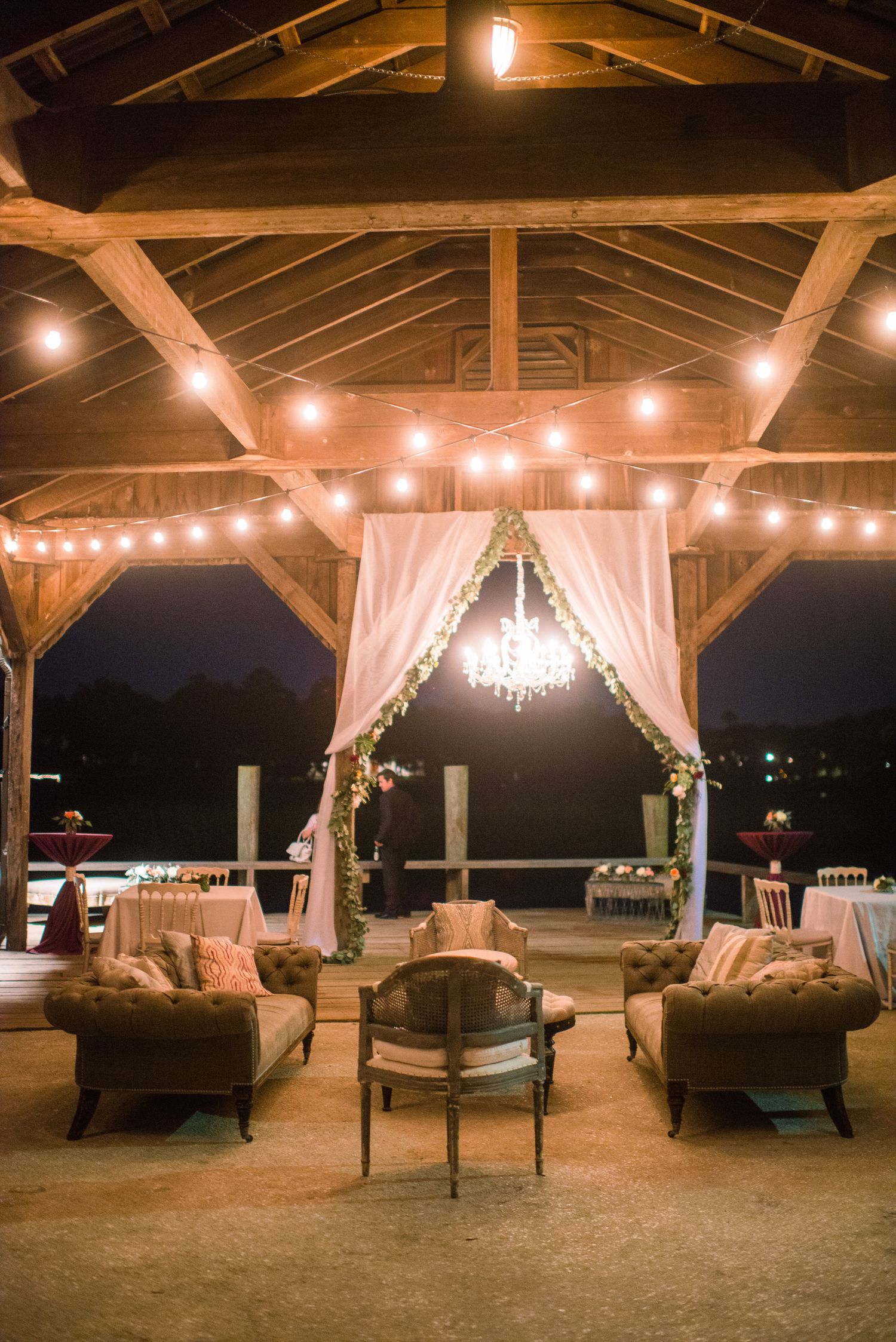 Elegant outdoor wedding lounge area with string lights, white drapes, and cozy seating under a wooden pavilion.