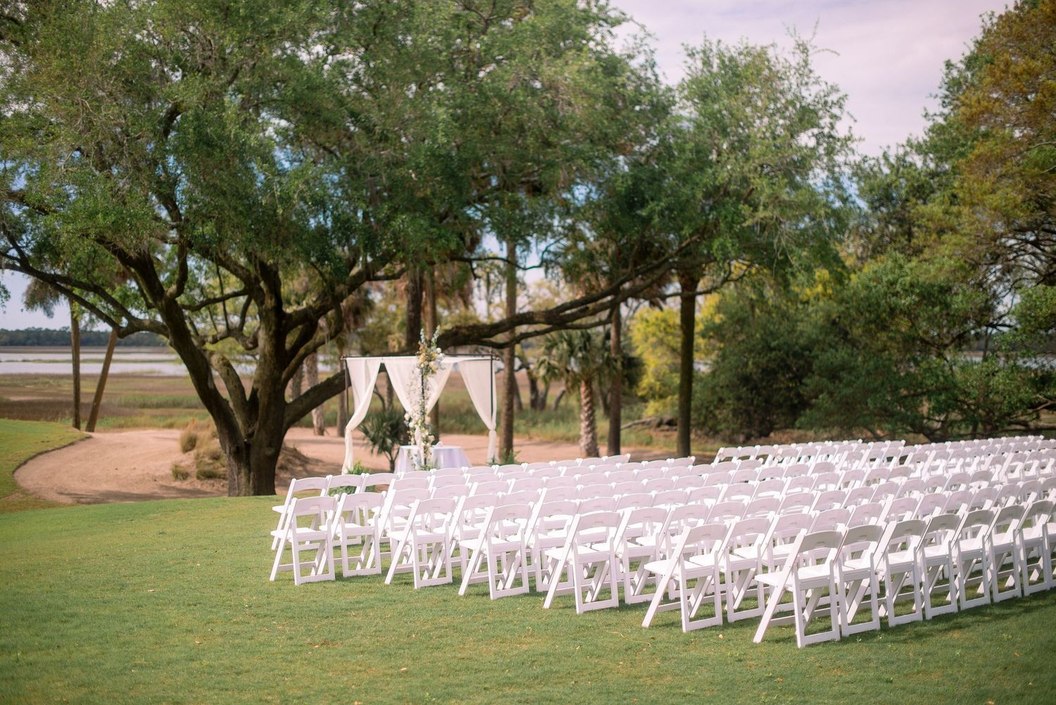 White wedding chairs arranged in rows under large oak trees for an outdoor ceremony at sunset.