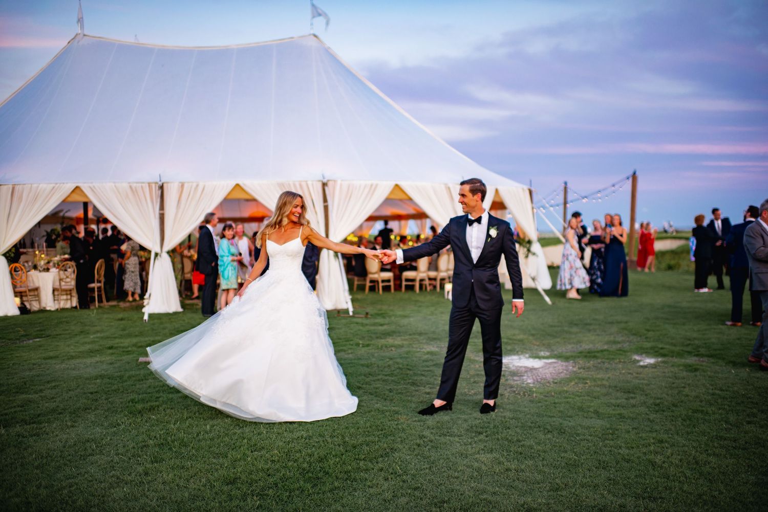A wedding party poses in front of white tents on a grassy field during sunset.