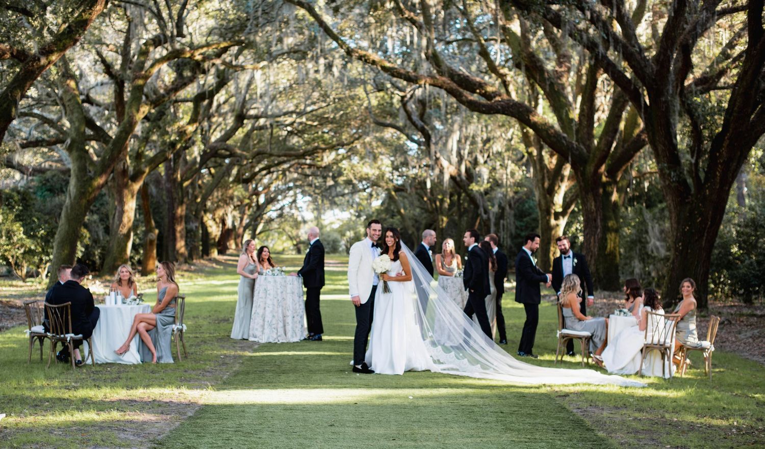 A wedding ceremony takes place under majestic oak trees draped with Spanish moss in a serene outdoor setting.